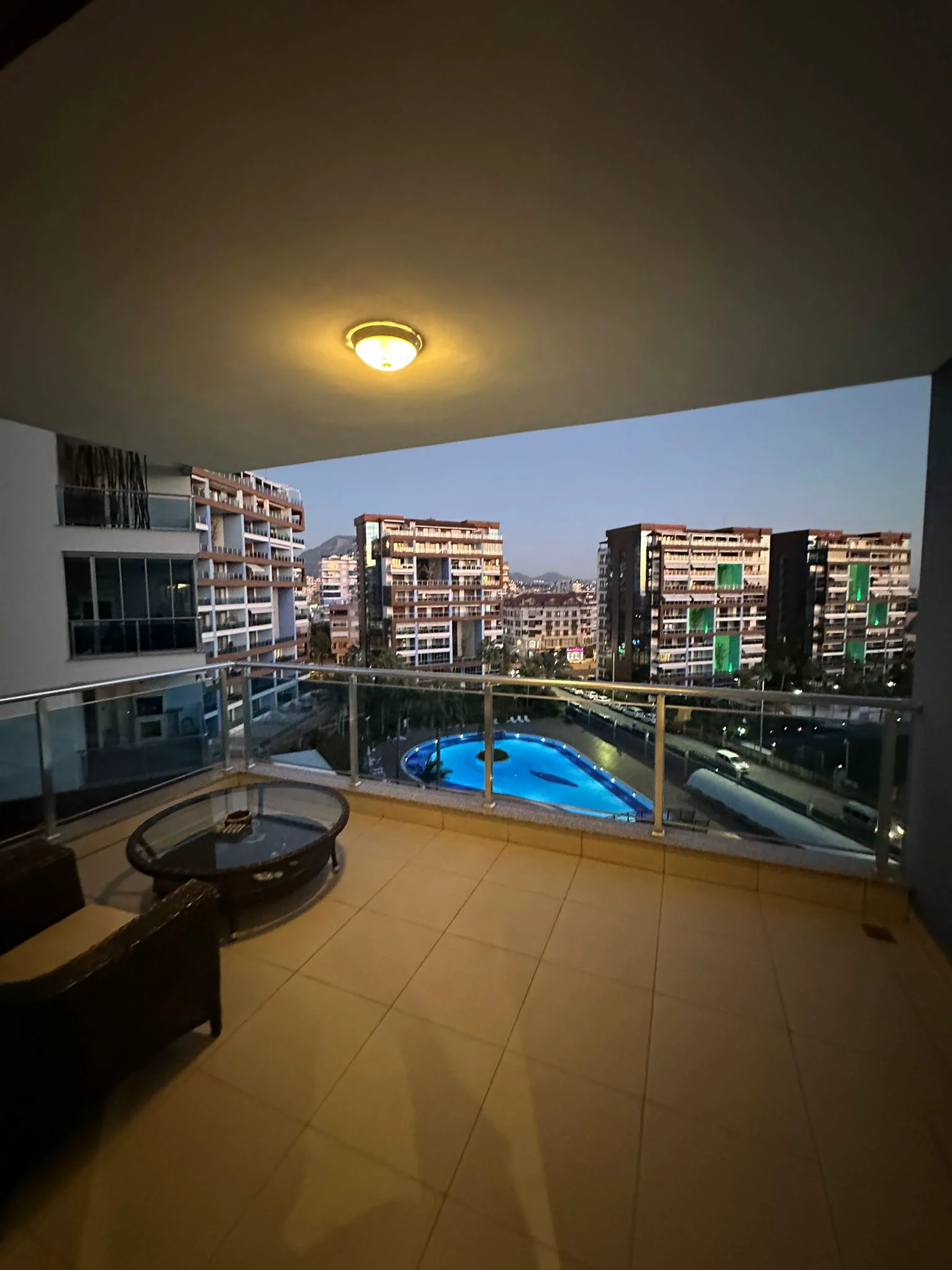 Moderner Balkon mit beleuchtetem Pool und Stadtblick. Moderner Apartment-Balkon bei Nacht mit hellen Fliesen und Glasgeländer, ausgestattet mit Außenmöbeln und Deckenbeleuchtung. Blick auf einen hell erleuchteten blauen Swimmingpool, moderne Wohngebäude und ferne Berge.