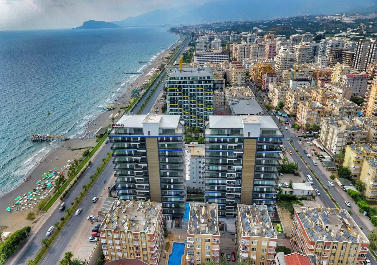 Panoramablick auf die Stadt am Meer mit modernen Apartmenttürmen. Luftaufnahme einer lebendigen türkischen Küstenstadt mit modernen Hochhauswohnungen, einem breiten Sandstrand mit Sonnenschirmen und dem tiefblauen Mittelmeer, das sich bis zu fernen Bergen erstreckt.
