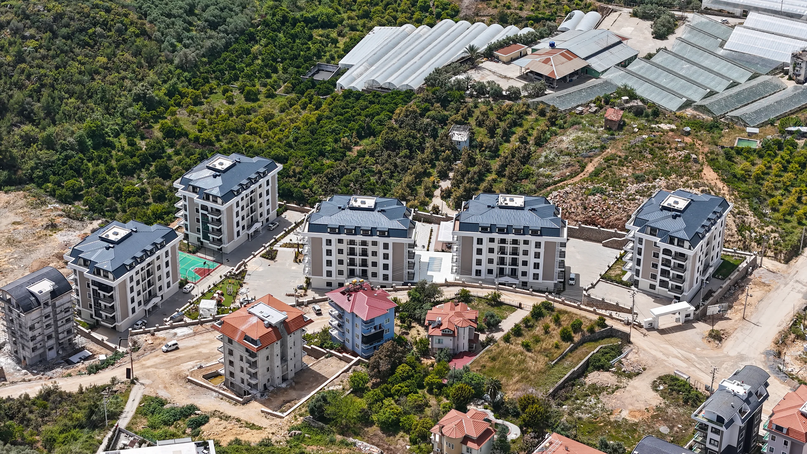 Moderner Wohnkomplex inmitten grüner Hügel und Landwirtschaft. Luftaufnahme eines neu entwickelten Wohnkomplexes mit mehreren modernen Apartmentgebäuden, hellen Fassaden und dunklen Dächern, vor dem Hintergrund üppig grüner Hügel und landwirtschaftlicher Gewächshäuser.
