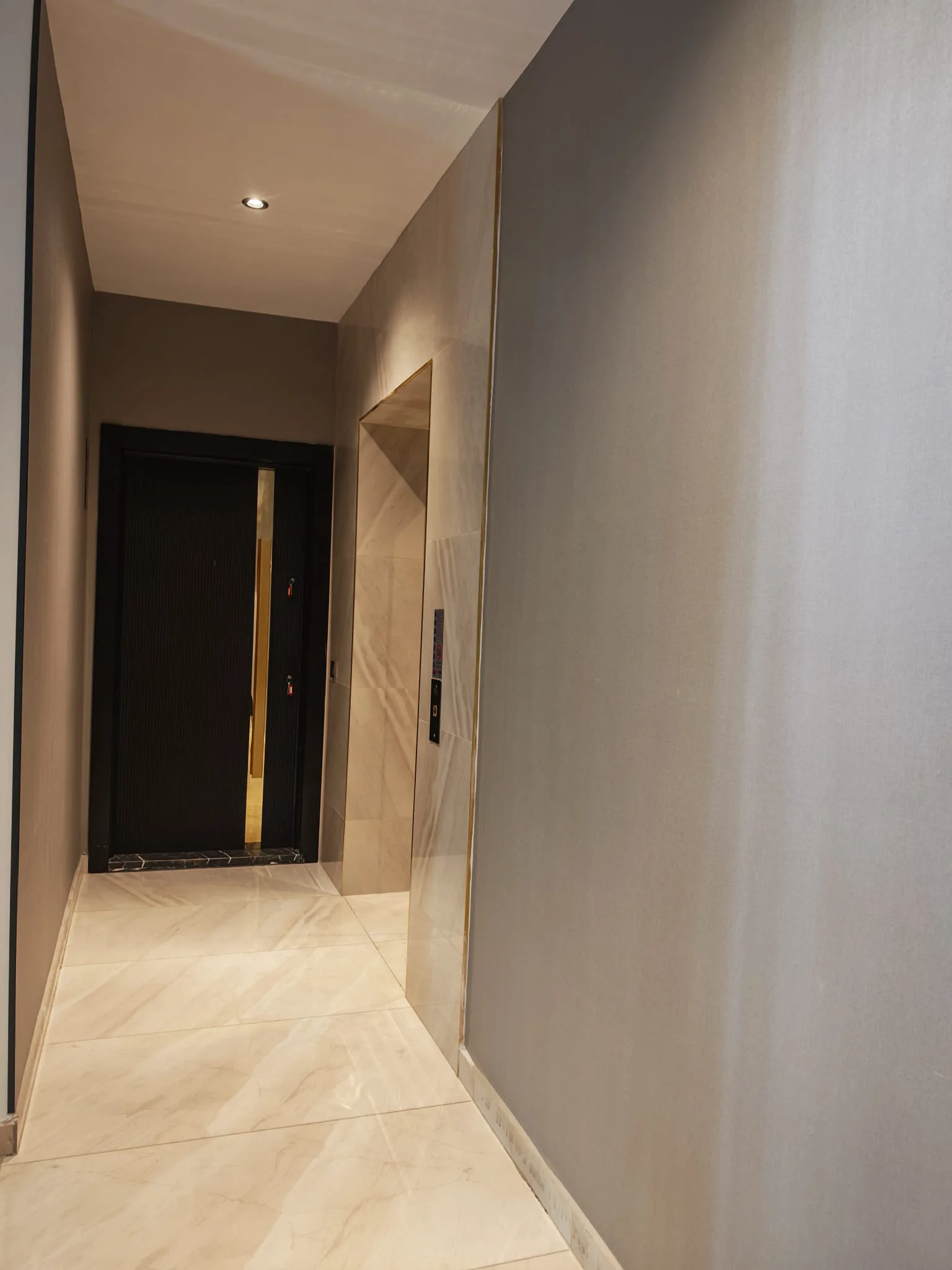 Moderner Flur in einem Apartmenthaus mit elegantem Aufzugszugang. Ein makelloser moderner Flur mit hellen Bodenfliesen in Marmoroptik, Deckeneinbauleuchten und einer dunklen Wohnungstür mit vertikalen Goldakzenten. Ein eleganter Aufzug mit digitalem Bedienfeld ist sichtbar.