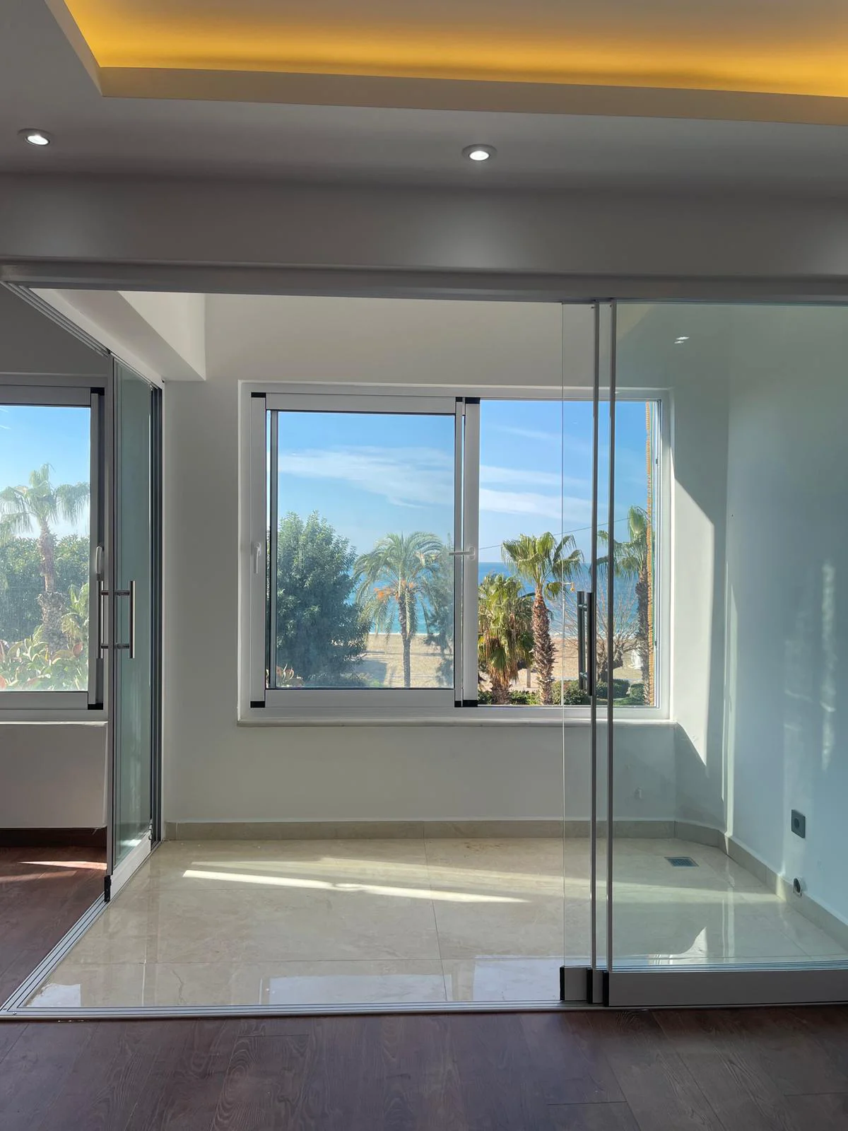 Helles modernes Zimmer mit Panorama-Blick auf Strand und Meer. Modernes Zimmer mit Blick auf einen Sandstrand mit Palmen und das blaue Meer. Ausgestattet mit polierten Fliesenböden, eleganten Glastüren und heller Einbaubeleuchtung.