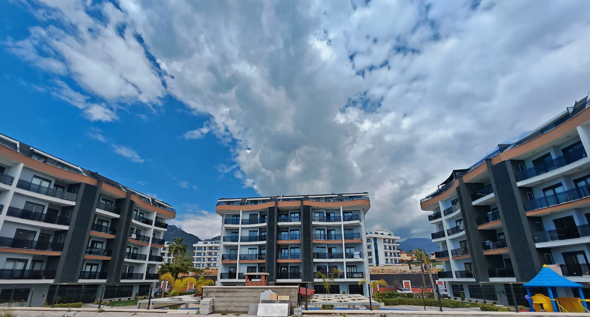 Moderner Wohnkomplex mit Balkonen und Bergblick. Außenansicht eines modernen Wohnkomplexes mit mehreren Wohngebäuden, die jeweils über zahlreiche Balkone verfügen. Beinhaltet einen Kinderspielplatz und Bergblick unter einem dynamischen Himmel.