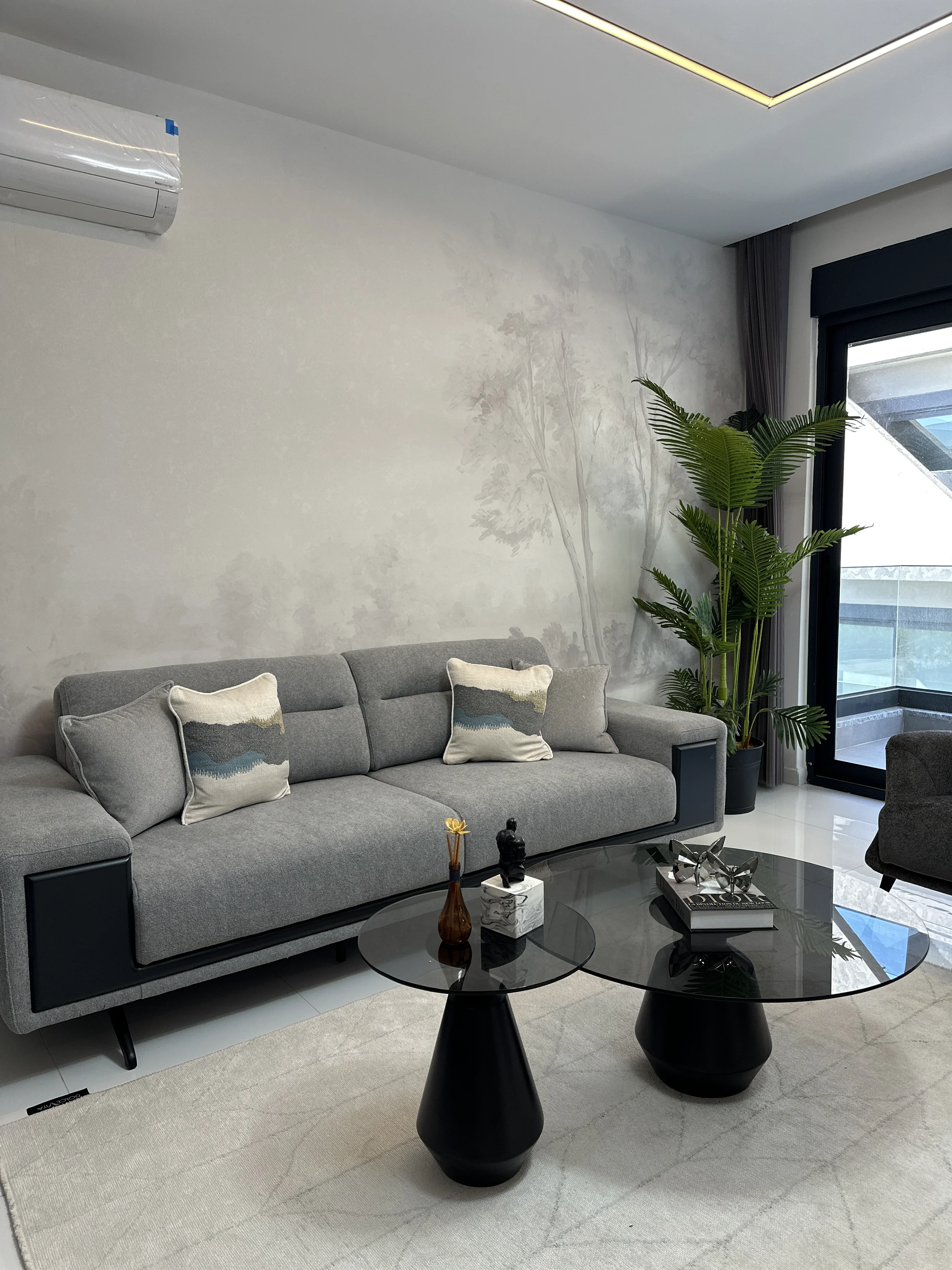 Modernes Wohnzimmer mit Baumwandbild und Balkonzugang. Ein helles, modernes Wohnzimmer mit einem bequemen grauen Sofa, einem einzigartigen Baumwandbild und eleganten Glas-Couchtischen. Es verfügt über weißen Fliesenboden, Klimaanlage und Balkonzugang.