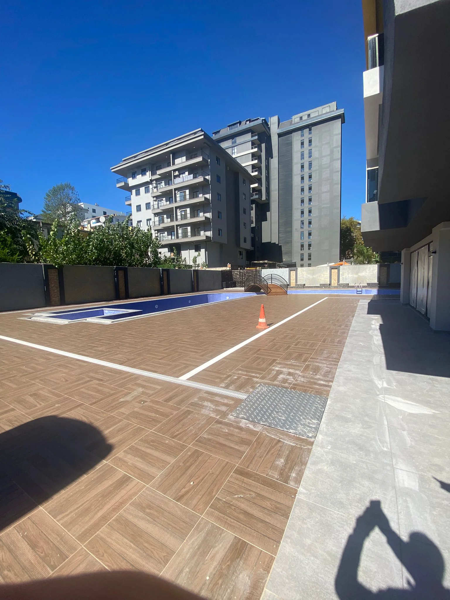 Großzügiger Außenpoolbereich Moderne Wohngebäude. Moderner Außenpoolbereich einer Wohnanlage mit großem Schwimmbecken und dekorativer Brücke. Mit neuem braun gefliesten Deck, klarem blauem Himmel und modernen grauen Gebäuden.