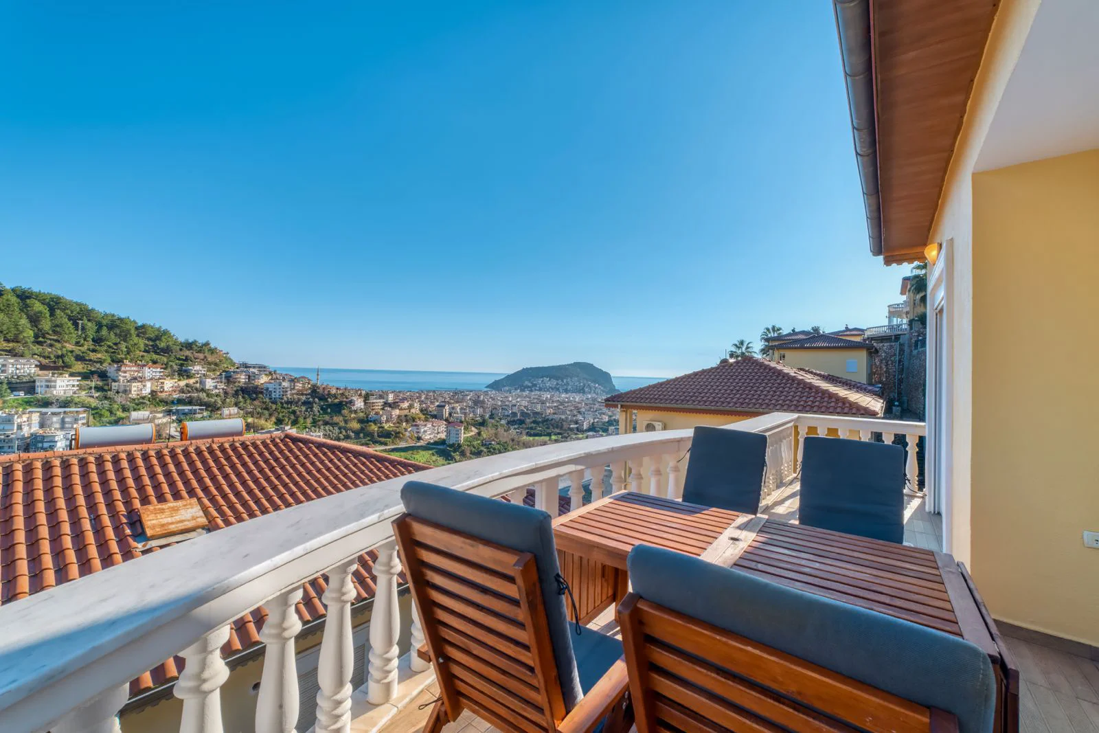 Panoramabalkon mit atemberaubendem Meer- und Stadtblick. Geräumiger, gepflegter Balkon mit einer Essgruppe aus Holz. Bietet einen Panoramablick auf eine Küstenstadt, rote Ziegeldächer und das tiefblaue Mittelmeer unter einem klaren, hellen Himmel.