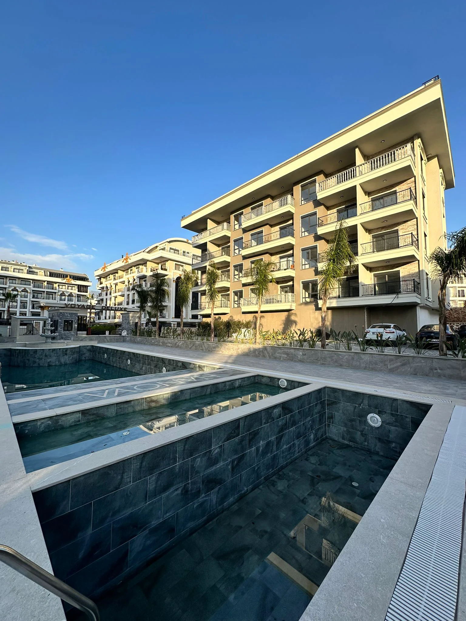 Außenpool einer modernen Wohnanlage. Außenansicht einer modernen Wohnanlage mit mehreren Außenpools mit dunklen Fliesen. Im Hintergrund sind neue Apartmentgebäude mit Balkonen und parkähnlich gestaltete Bereiche mit Palmen zu sehen.