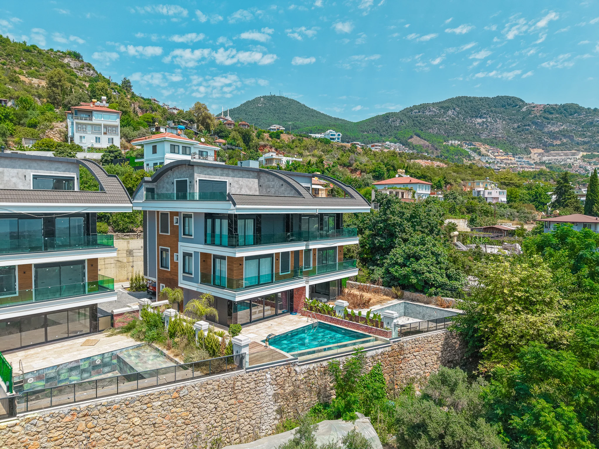 Moderne Hangvillen mit privaten Pools und Bergblick. Außenansicht moderner, luxuriöser Hangvillen mit mehrstöckigem Design, großen Glasbalkonen und privaten Swimmingpools vor der Kulisse grüner Berge unter klarem Himmel.