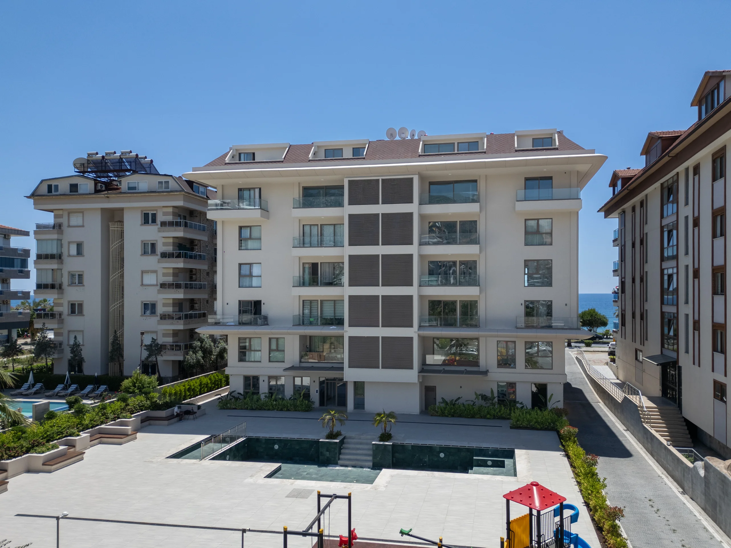Modernes Apartmentgebäude mit Pool und Meerblick. Außenansicht eines modernen Apartmentgebäudes mit Swimmingpool, Kinderspielplatz und angelegten Gärten. Die gepflegte Anlage bietet mehrere Balkone und einen klaren Meerblick.