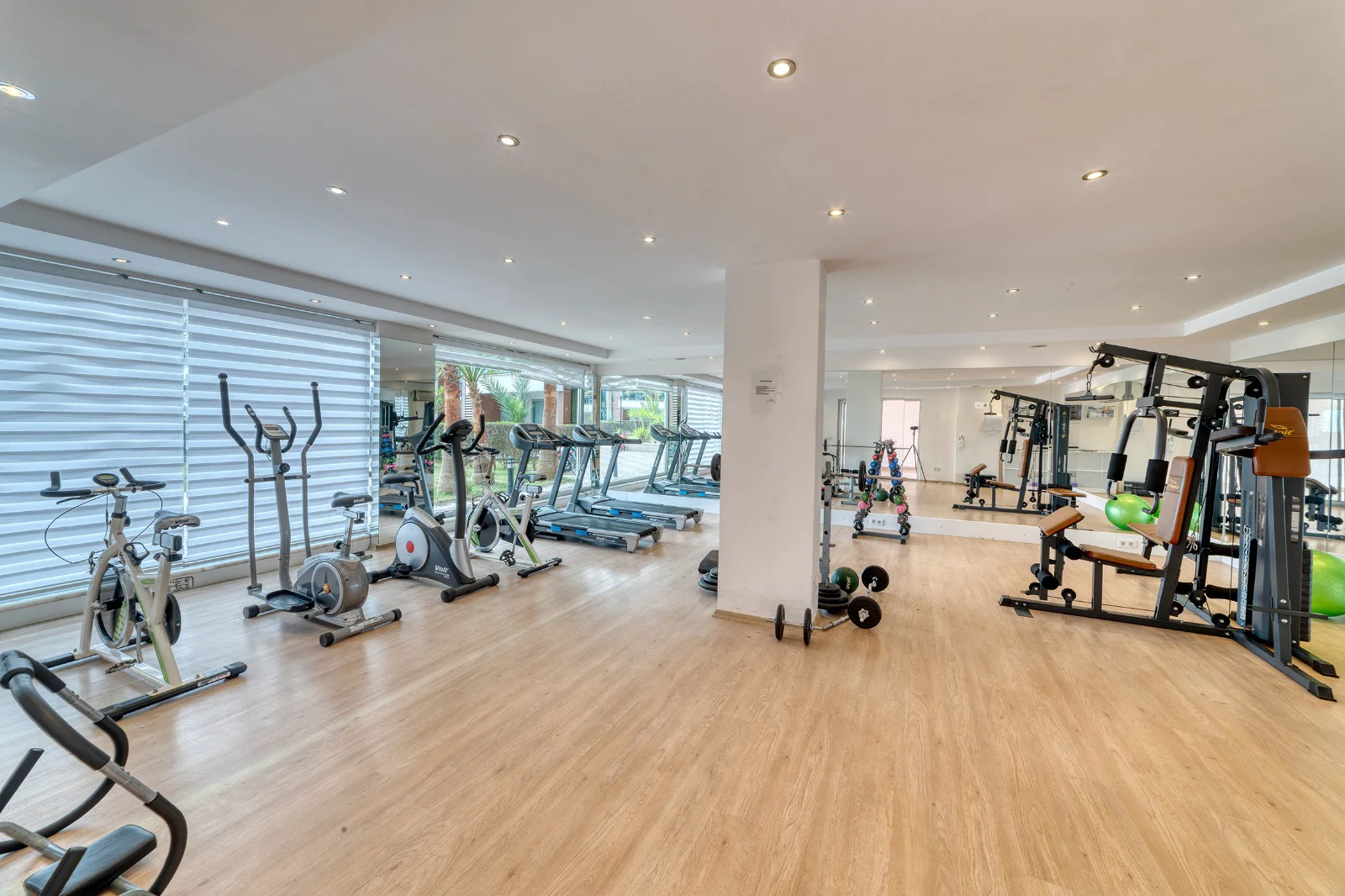 Modernes Anwesen-Fitnessstudio mit umfangreicher Ausrüstung und Tageslicht. Ein modernes und gut gepflegtes Fitnessstudio mit hellem Holzboden, viel Tageslicht durch große Fenster mit Jalousien und einer umfassenden Auswahl an Fitnessgeräten, darunter Laufbänder, Fahrräder und Kraftmaschinen.