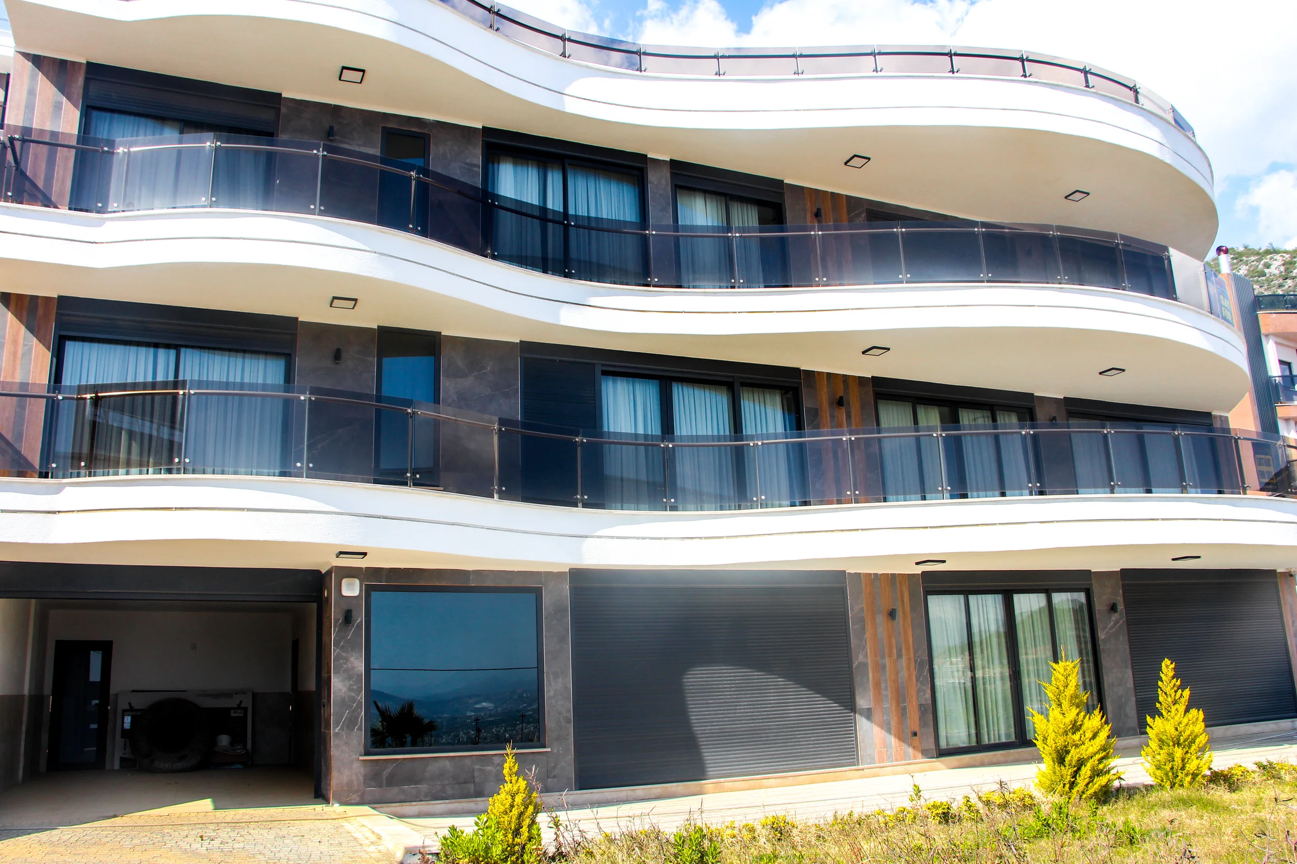 Moderne Außenansicht eines Apartmenthauses mit stilvollen Balkonen. Außenansicht eines modernen Wohngebäudes mit eleganten, geschwungenen weißen Balkonen, dunklen Glasbrüstungen und großen Fenstern. Elegante dunkle Fassade mit Holzakzenten. Parkhauseinfahrt im Erdgeschoss sichtbar.