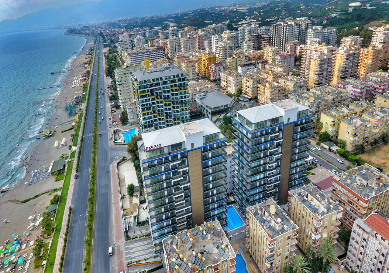 Luftaufnahme: Moderne Meerblick-Apartments mit Stadtstrand. Eine weitläufige Luftaufnahme einer lebhaften türkischen Küstenstadt, die moderne Hochhaus-Wohngebäude mit Schwimmbädern, einen langen Sandstrand und das funkelnde Meer zeigt.