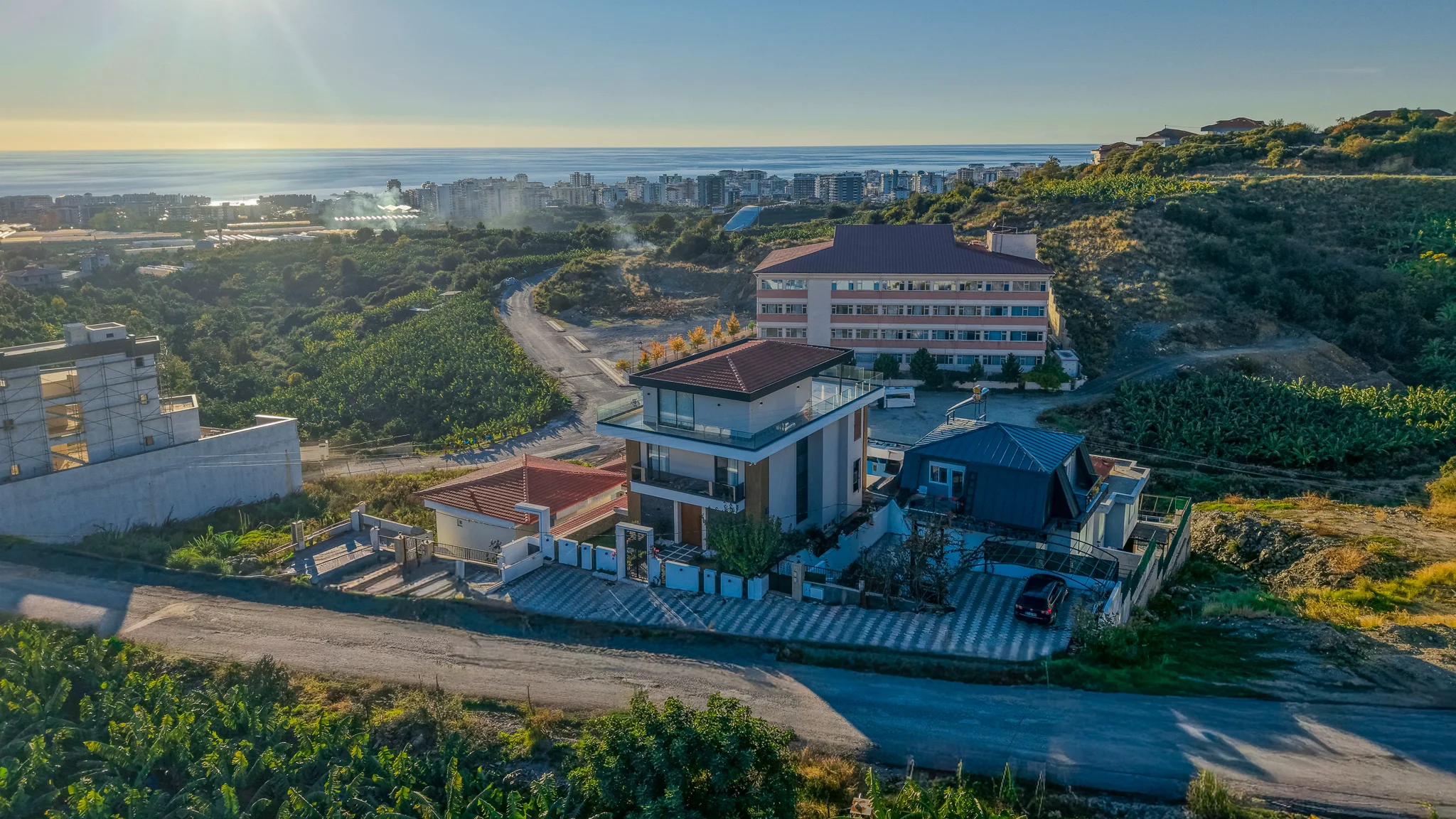 Moderne Villa mit Panoramablick auf Meer und Stadt. Luftaufnahme einer modernen mehrstöckigen Villa an einem grünen Hang. Mit Dachterrasse und Balkonen mit Panoramablick auf die ferne Stadt und das glitzernde blaue Meer.
