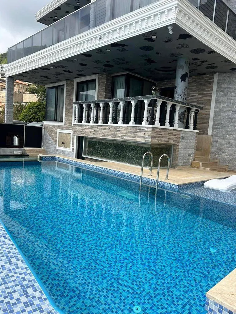 Außenansicht einer Luxusvilla mit privatem Swimmingpool und Balkon. Außenansicht einer luxuriösen modernen Villa mit einem großen, glitzernden Außenpool mit blauen Mosaikfliesen. Ein klassischer Balkon ist darüber sichtbar und unterstreicht das opulente Design.