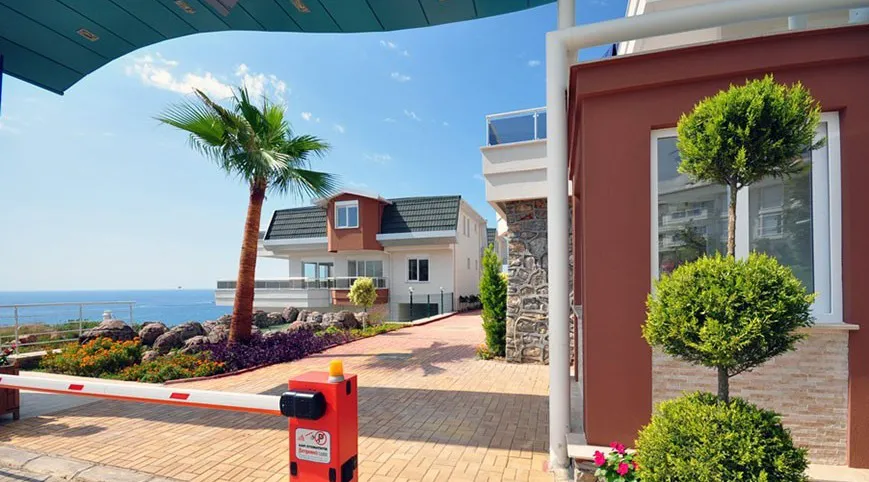 Bewachter Eingang zu Luxusvillen mit Meerblick. Außenansicht einer gesicherten, bewachten Wohnanlage mit einer rot-weißen Schranke, Palmen und modernen Villen mit Blick auf das Mittelmeer unter einem klaren blauen Himmel.
