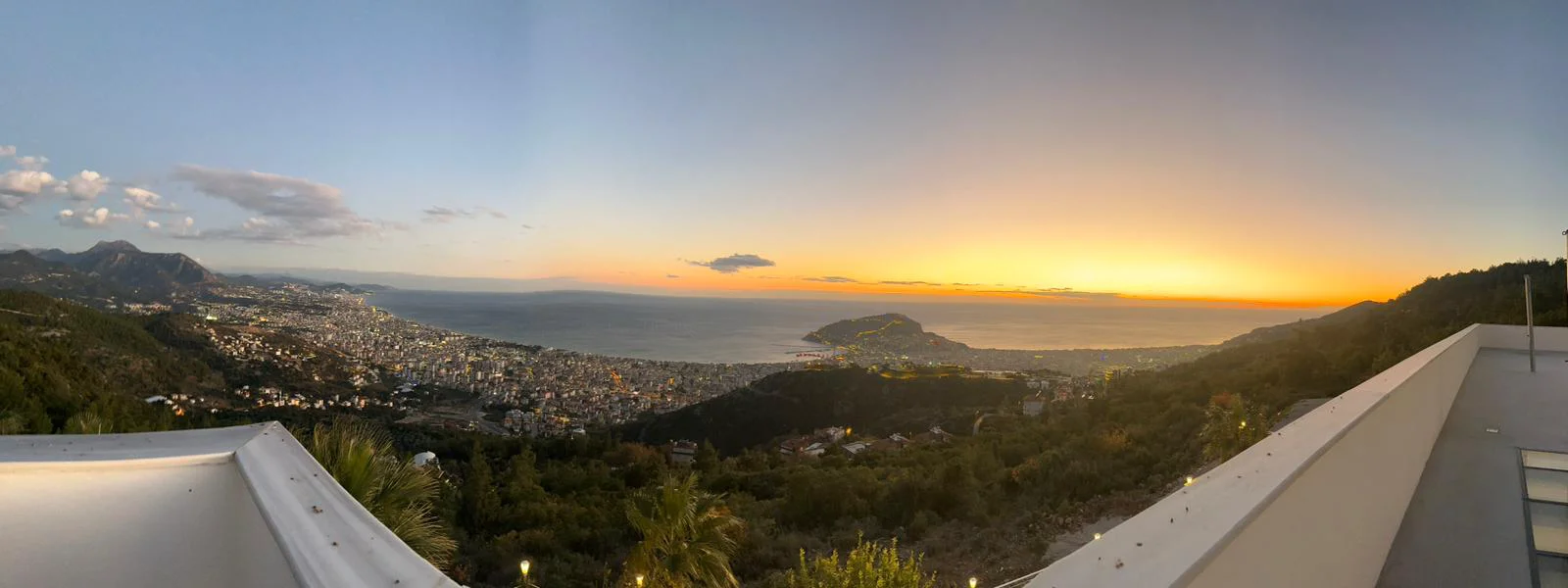 Spektakulärer Panorama-Meer- und Stadtblick bei Sonnenuntergang. Ein Panoramablick von einer modernen Terrasse bei Sonnenuntergang, der eine geschäftige Stadt zwischen Bergen und dem ruhigen Mittelmeer zeigt, mit einem leuchtend orangefarbenen und gelben Himmel.