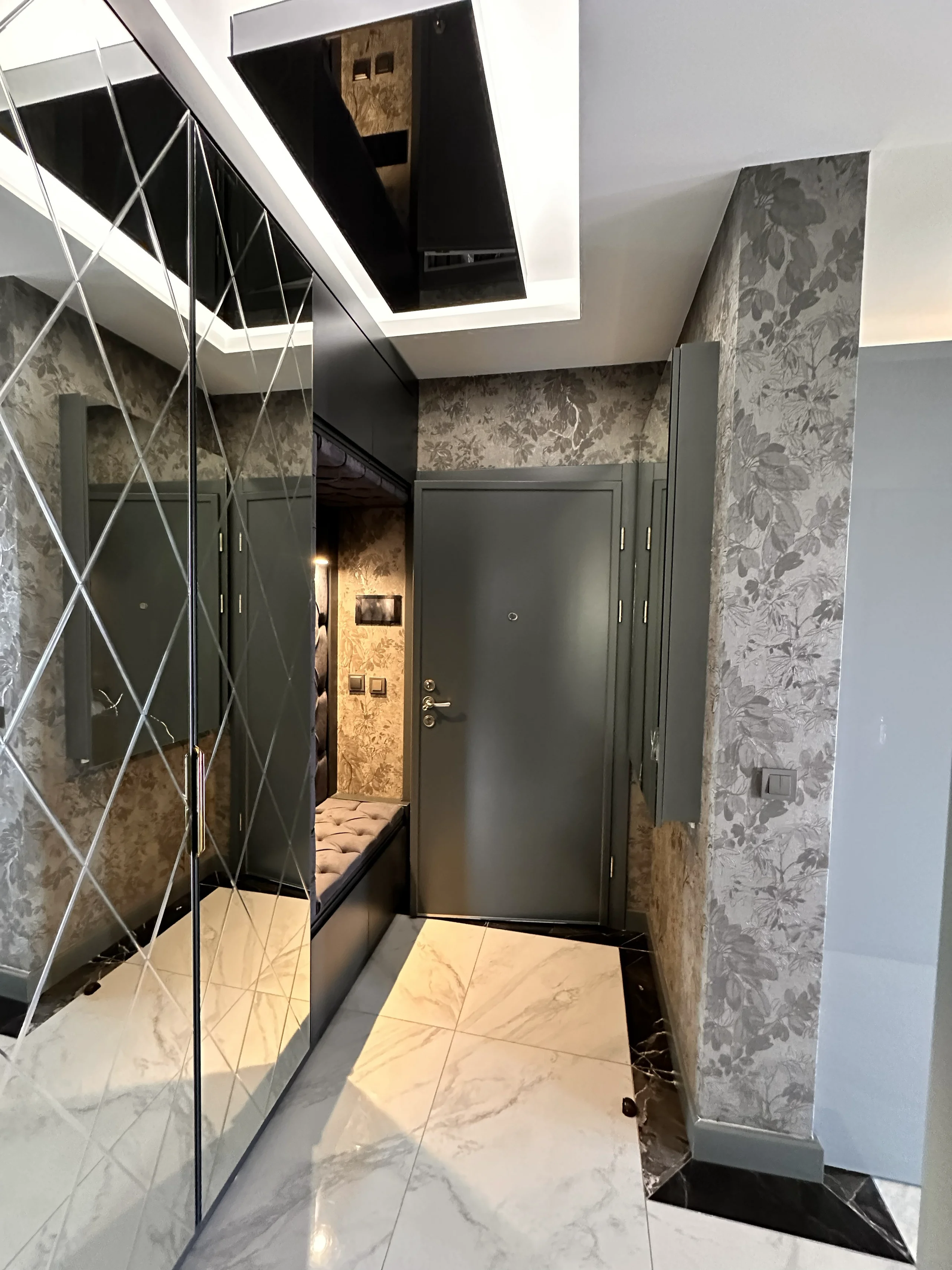 Stilvoller moderner Eingangsflur mit Spiegelgarderobe. Moderner Eingangsflur mit weißen Fliesen in Marmoroptik, eleganter grauer Mustertapete und einer großen Spiegelgarderobe. Mit einer dunkelgrauen Tür und einer integrierten Sitzbank.
