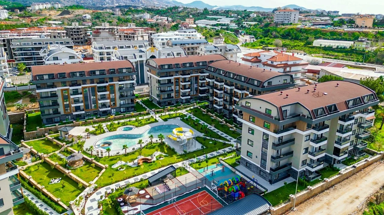 Moderner Apartmentkomplex mit umfangreicher Ausstattung und Pool. Luftaufnahme einer modernen Wohnanlage mit mehreren Gebäuden, einem großen Außenpool mit Wasserrutsche, grünen Gärten, einem Spielplatz und einem Sportplatz.