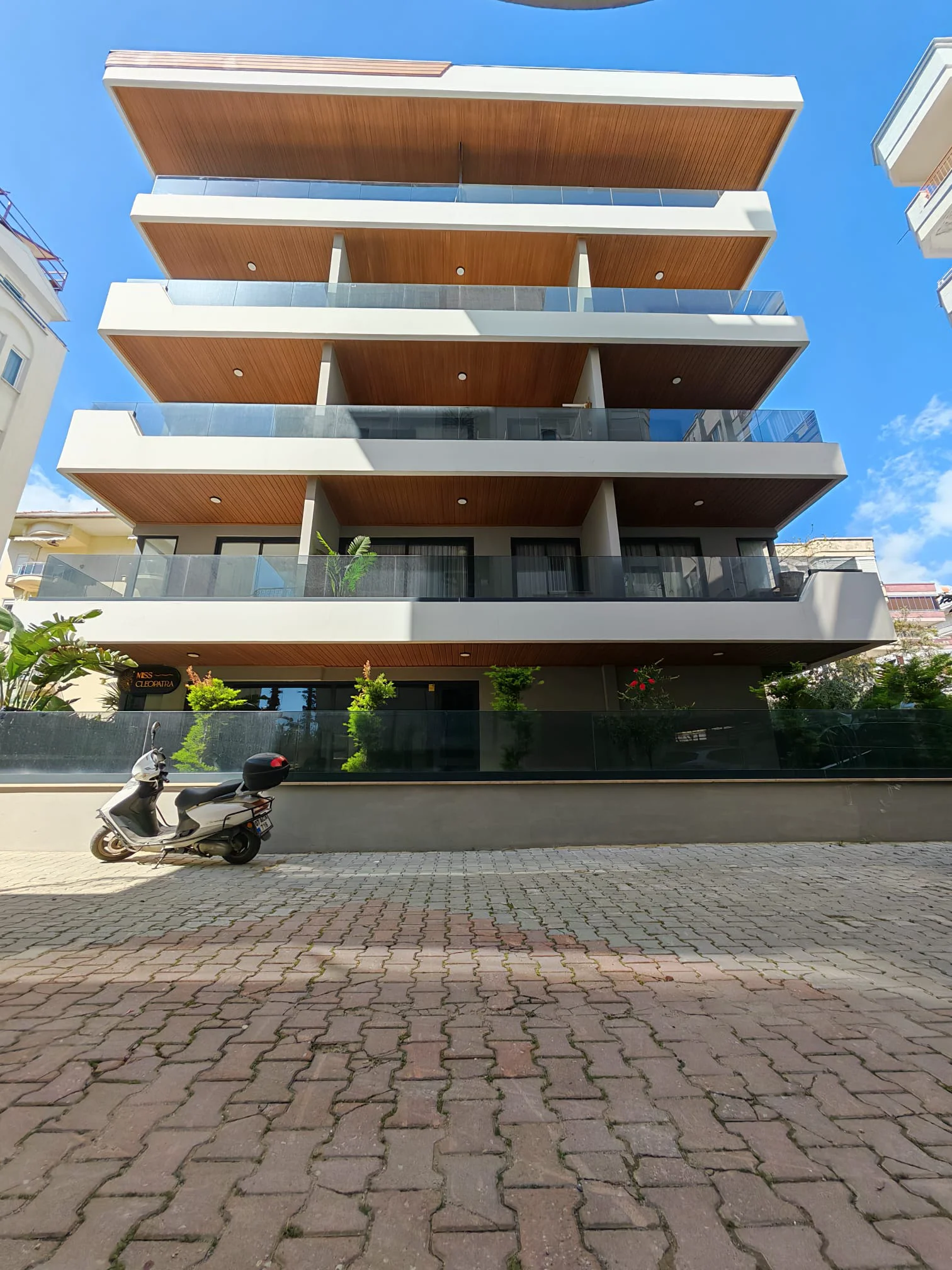 Modernes mehrstöckiges Wohngebäude mit Balkonen. Außenansicht eines zeitgenössischen mehrstöckigen Wohnhauses mit modernem Design, Glasbalkonen mit Holzdecken und üppiger Bepflanzung im Erdgeschoss. Ein Roller parkt auf dem gepflasterten Vorplatz.