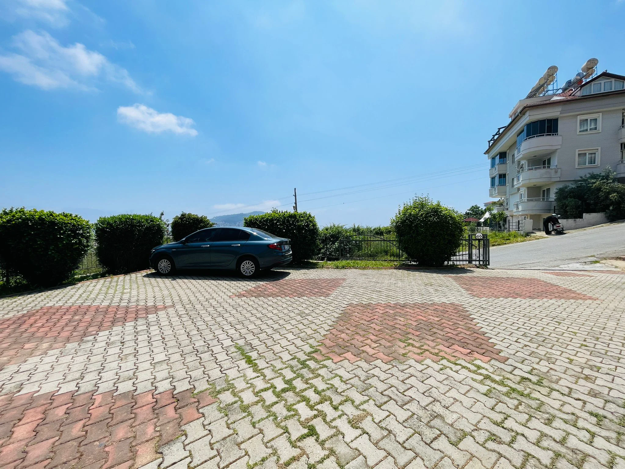 Gepflasterter Außenparkplatz mit Blick auf ein Mehrfamilienhaus. Gepflasterter Außenparkplatz mit einem Auto, umgeben von grünen Hecken. Im Hintergrund ein mehrstöckiges Wohngebäude mit Balkonen unter klarem Himmel.