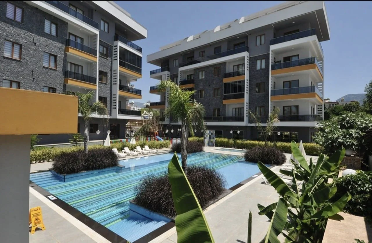 Moderner Apartmentkomplex mit glitzerndem Außenpool. Ein einladender Außenpoolbereich in einem modernen Apartmentkomplex. Mit einem großen, gefliesten Pool mit Sonnenliegen, umgeben von tropischen Pflanzen und zeitgenössischen Gebäuden.