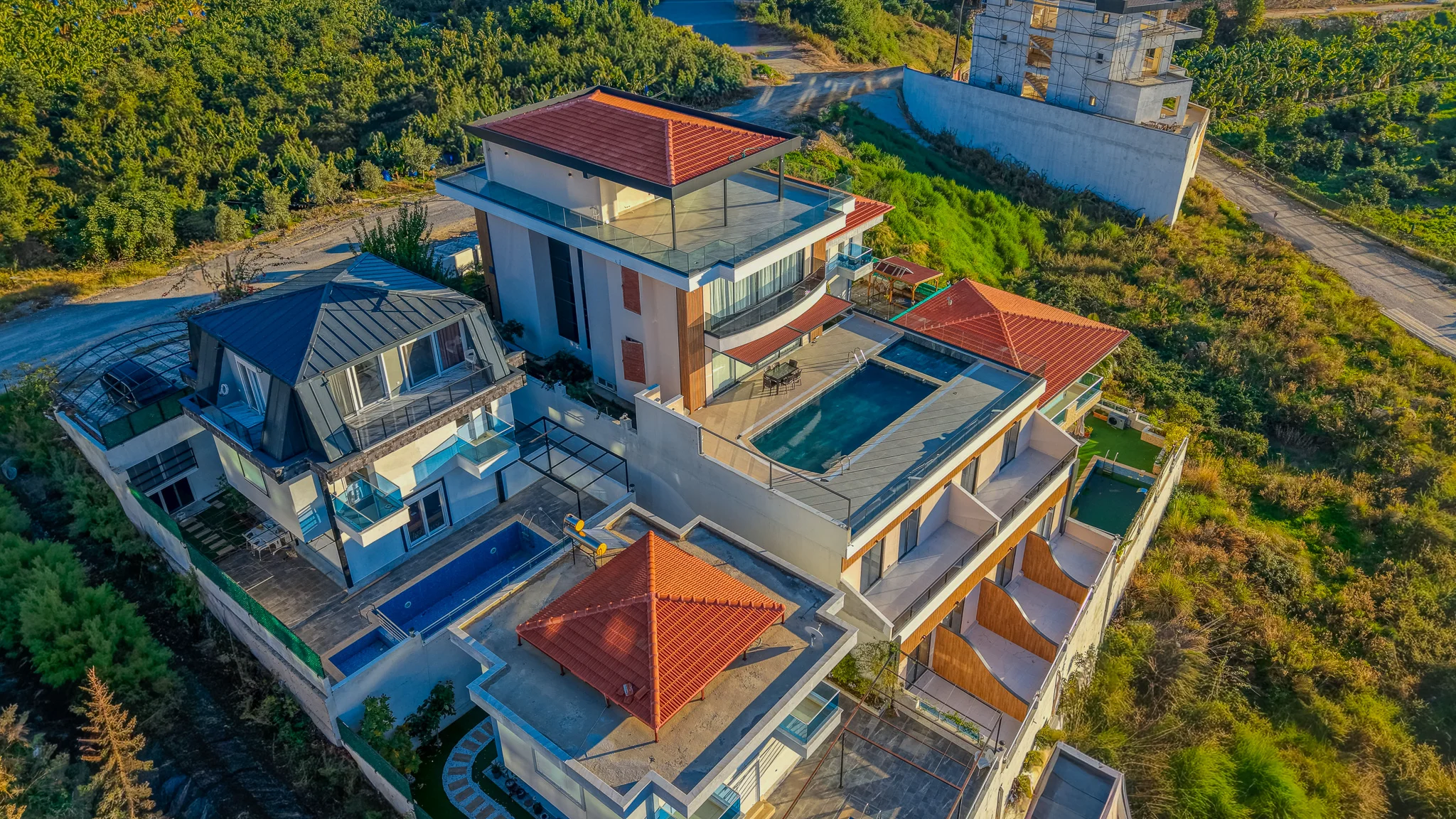 Luxuriöse Hangvillen mit privaten Pools und Panoramablick. Luftaufnahme eines modernen Immobilienkomplexes am Hang mit mehreren Luxusvillen, privaten Außenpools, geräumigen Balkonen mit Glasgeländer und terrassierten Gärten inmitten von üppigem Grün.