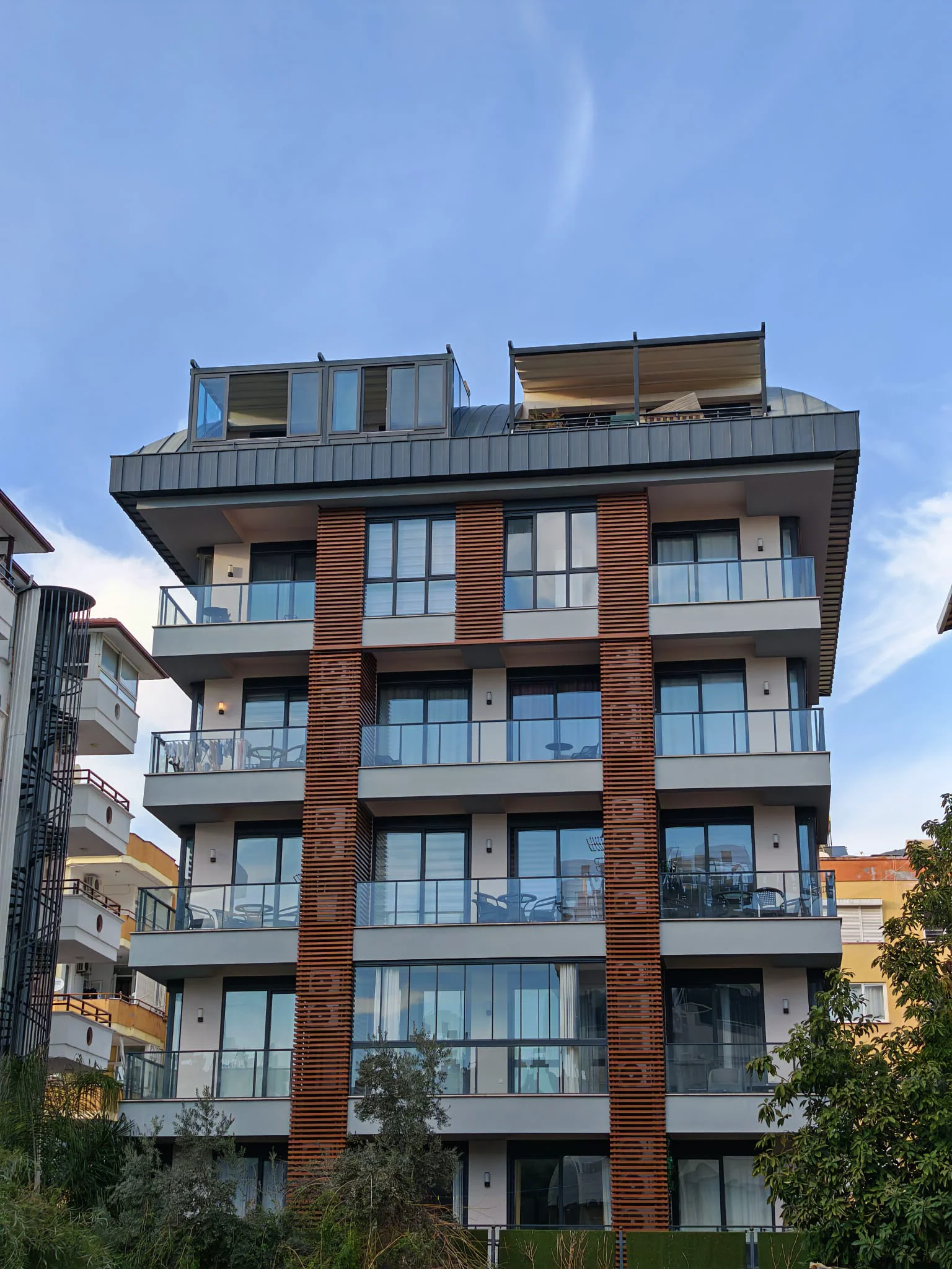 Außenansicht eines modernen Mehrfamilienhauses mit Balkonen. Außenansicht eines modernen Wohngebäudes mit mehreren Balkonen, Glasgeländern und dekorativen Holzfassadenelementen. Das zeitgenössische Design umfasst große Fenster und eine Dachterrasse mit ausfahrbarer Markise.