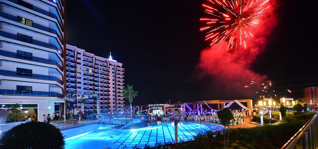 Lebendiges Nachtleben am Resort-Pool mit Feuerwerk. Lebhafte Nachtansicht einer modernen Wohnanlage mit einem beleuchteten Swimmingpool, einem aktiven Veranstaltungsbereich im Freien und rotem Feuerwerk, das den dunklen Himmel erhellt.