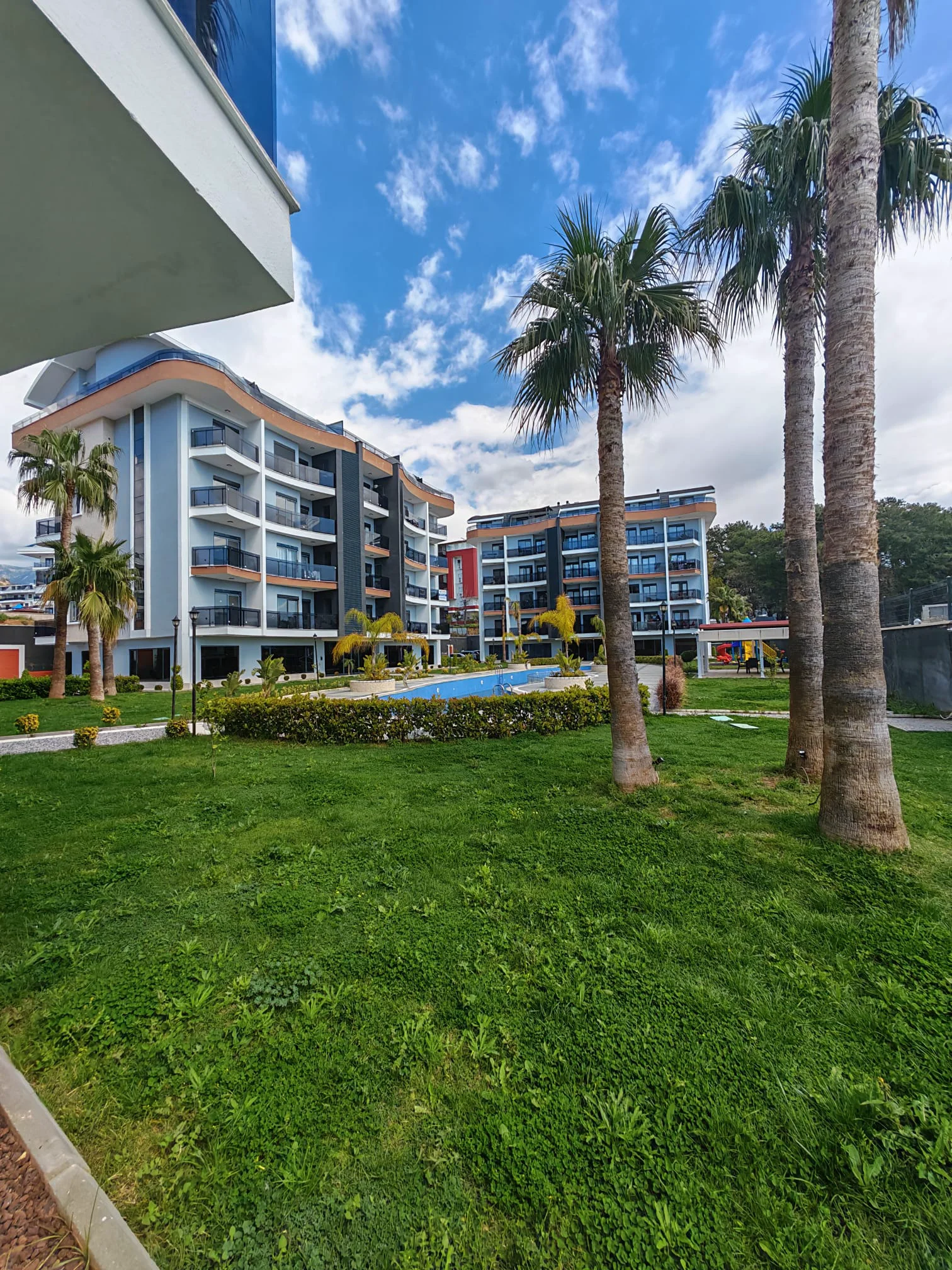 Moderne Apartmentanlage mit Pool und üppigen Gärten. Außenansicht einer modernen Wohnanlage mit mehrstöckigen Gebäuden mit Balkonen, einem Swimmingpool und üppigen grünen Gärten mit hohen Palmen.