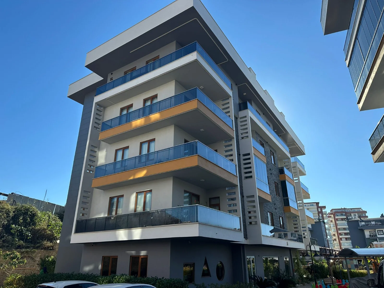 Modernes Apartmentgebäude mit stilvollen Balkonen. Außenansicht eines modernen mehrstöckigen Wohngebäudes unter klarem blauem Himmel. Es verfügt über eine stilvolle grau-weiße Fassade mit zahlreichen Balkonen, die mit blauen Glasgeländern und orangefarbenen Akzenten ausgestattet sind.