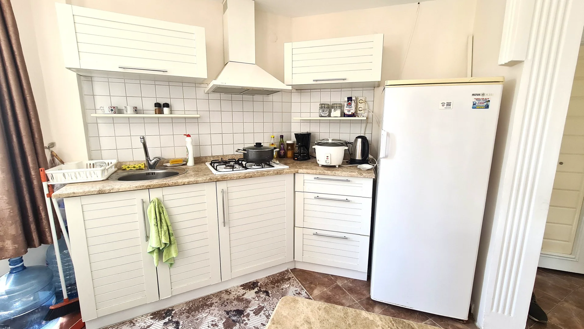 Funktionale kompakte Küche mit weißen Schränken und Geräten. Eine kompakte moderne Küche mit weißen Lamellenschränken, Fliesenspiegel und einer hellen Arbeitsplatte. Zur Ausstattung gehören ein Gasherd, eine Spüle, ein Kühlschrank und verschiedene Kleingeräte.