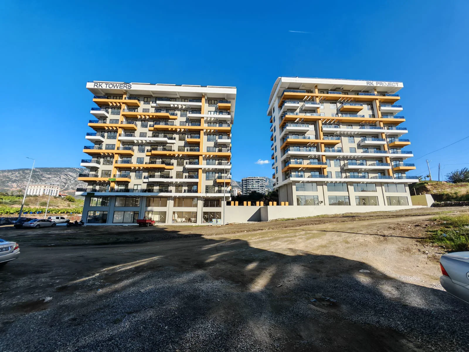 Moderne Außenansicht der Wohngebäude RK Towers und RK Project. Außenansicht von zwei modernen, neu gebauten Wohnhochhäusern, RK TOWERS und RK PROJECT, mit zahlreichen Balkonen mit Glasgeländern, Gewerbeflächen im Erdgeschoss und strahlend blauem Himmel.