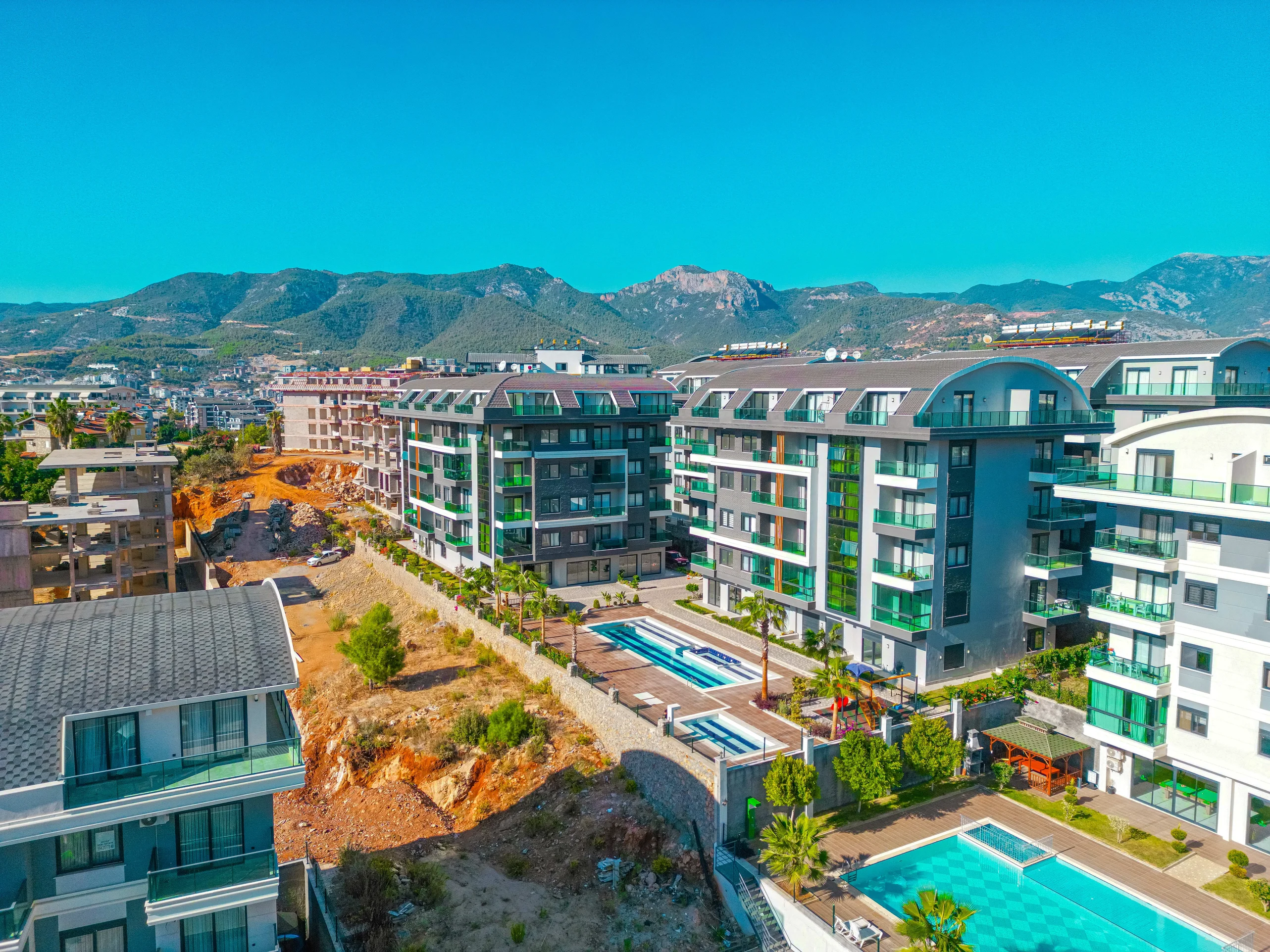 Moderner Wohnkomplex mit mehreren Pools und Bergblick. Luftaufnahme eines modernen Wohnkomplexes mit mehreren Außenpools, gepflegten Gärten und zeitgenössischen Gebäuden vor einer Bergkulisse unter klarem blauem Himmel.