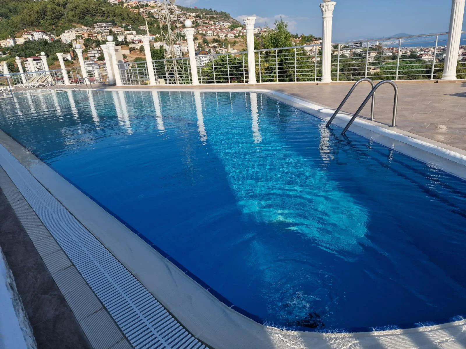 Außenpool mit Panorama-Stadt- und Meerblick. Ein einladender Außenpool mit klarem blauem Wasser und einer Metallleiter. Das moderne Pooldeck bietet einen atemberaubenden Panoramablick auf eine Stadt am Hang und das glitzernde Meer in der Ferne.