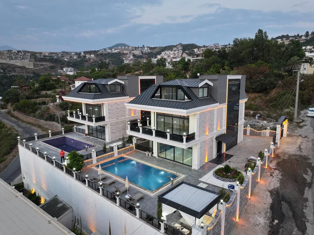 Moderne Doppelvillen mit privaten Pools und atemberaubendem Blick auf die Hügel. Außenansicht von zwei modernen, mehrstöckigen Villen, jeweils mit privatem Pool, Sonnenliegen und Balkonen, an einem terrassierten Hang mit malerischer Aussicht.