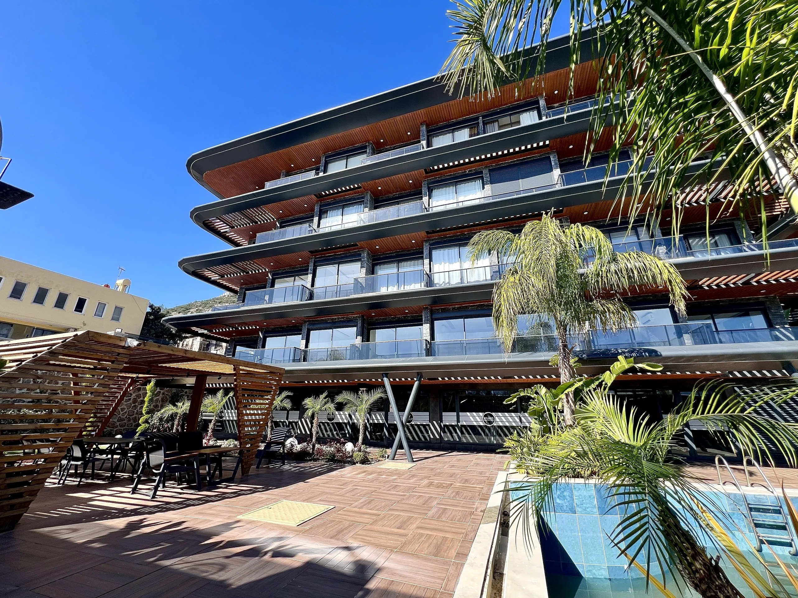 Modernes Apartmentgebäude mit Pool und Außenloungebereich. Außenansicht eines modernen mehrstöckigen Apartmentgebäudes mit weitläufigen Balkonen, einem schicken Außenloungebereich mit Sitzgelegenheiten in einem Holzpavillon, einem erfrischenden Swimmingpool und lebendigen Palmen.