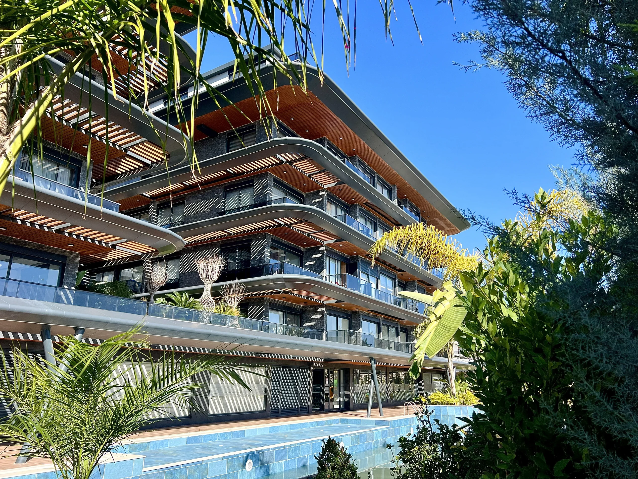 Modernes Luxus-Apartmentgebäude mit Pool und tropischem Garten. Außenansicht eines modernen mehrstöckigen Apartmentgebäudes mit geschwungenen Balkonen, Holzdetails und Glasgeländern. Ein blauer Swimmingpool und üppiges tropisches Grün schaffen einen luxuriösen Außenbereich.
