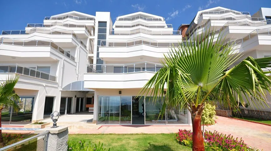 Moderne Außenansicht eines Apartmenthauses mit terrassierten Balkonen. Moderne weiße Außenfassade eines Apartmenthauses mit terrassierten Balkonen und Glasgeländern. Üppiger grüner Garten mit einer Palme und leuchtenden Blumen im Vordergrund unter blauem Himmel.