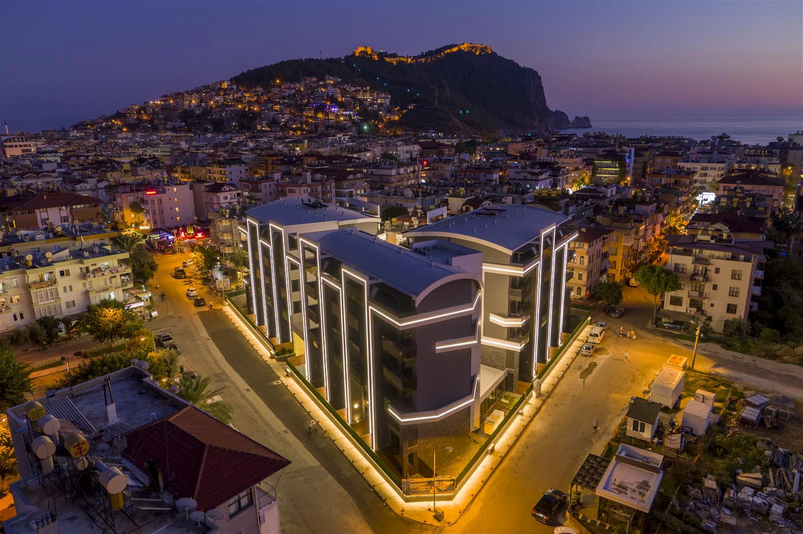 Modernes Apartmentgebäude mit Panoramablick auf Stadt und Meer in der Abenddämmerung. Eine Luftaufnahme eines modernen, beleuchteten Wohngebäudes in der Abenddämmerung, das seine elegante Fassade inmitten der Stadtlichter zeigt, mit dem Meer und der Burg von Alanya im Hintergrund.