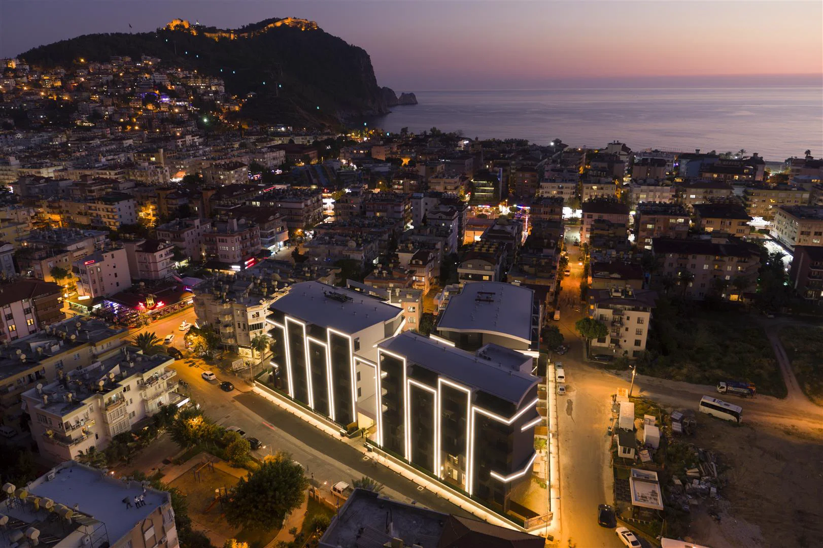 Moderne Wohngebäude mit atemberaubendem Blick auf die Stadt bei Nacht und das Meer. Eine nächtliche Luftaufnahme moderner Wohngebäude mit beleuchteten Fassaden, eingebettet in eine lebendige Stadt mit Blick auf das Meer und eine ferne Gebirgslandschaft.