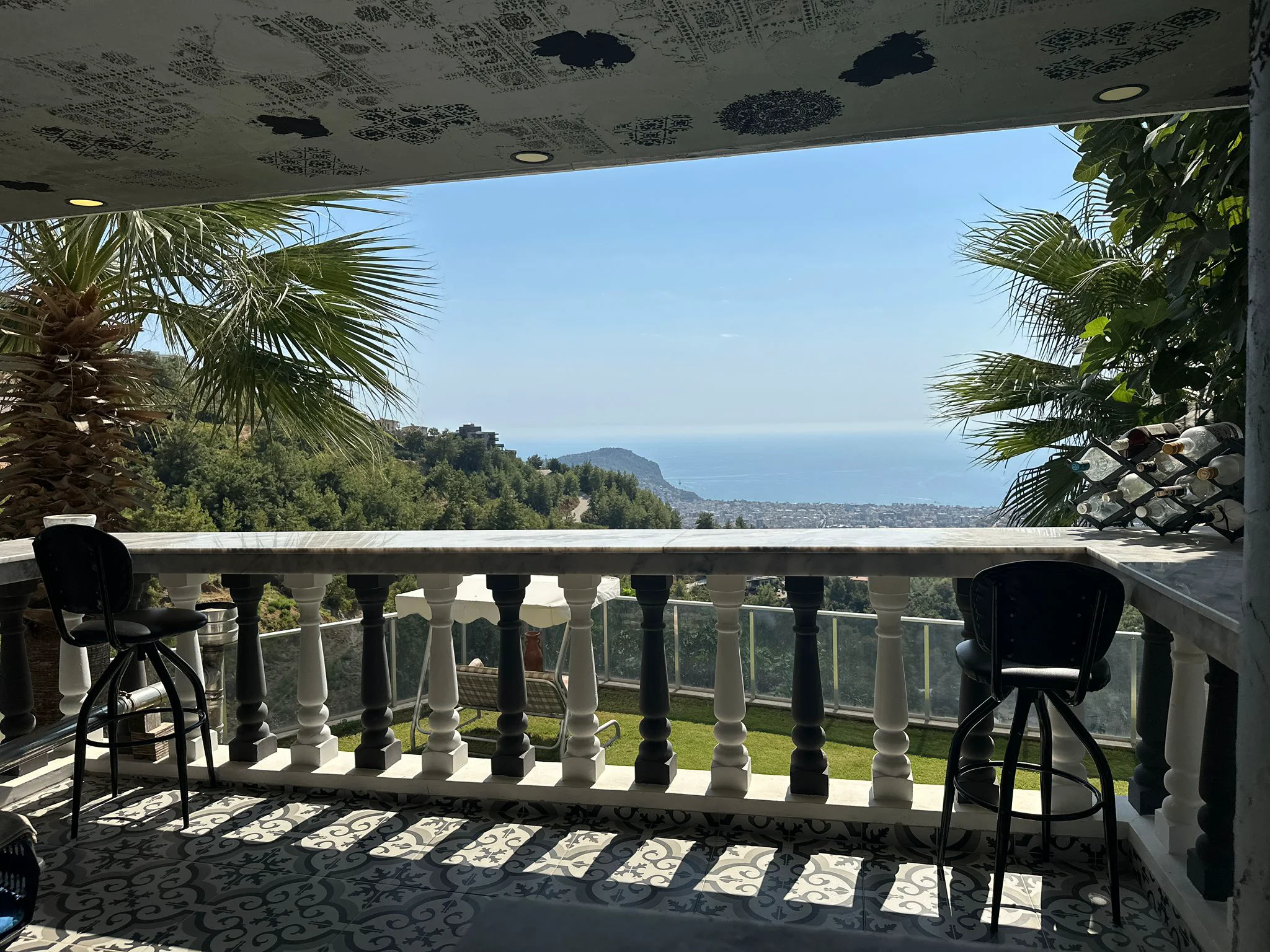 Großzügiger Balkon mit Panorama-Meer- und Bergblick. Ein geräumiger überdachter Balkon mit gemustertem Fliesenboden und zwei Barhockern, der einen atemberaubenden Panoramablick auf das Mittelmeer, die Küstenstadt und die grünen Berge bietet.