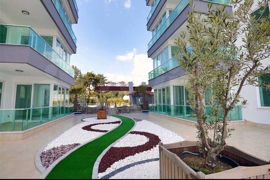 Moderner Apartmentgarten mit dekorativer Landschaftsgestaltung. Außenansicht einer modernen Wohnanlage mit einem schönen Innenhofgarten. Er verfügt über weiße und rote Dekosteine, einen grünen Pfad und einen Olivenbaum. Die Gebäude haben Glasbalkone.