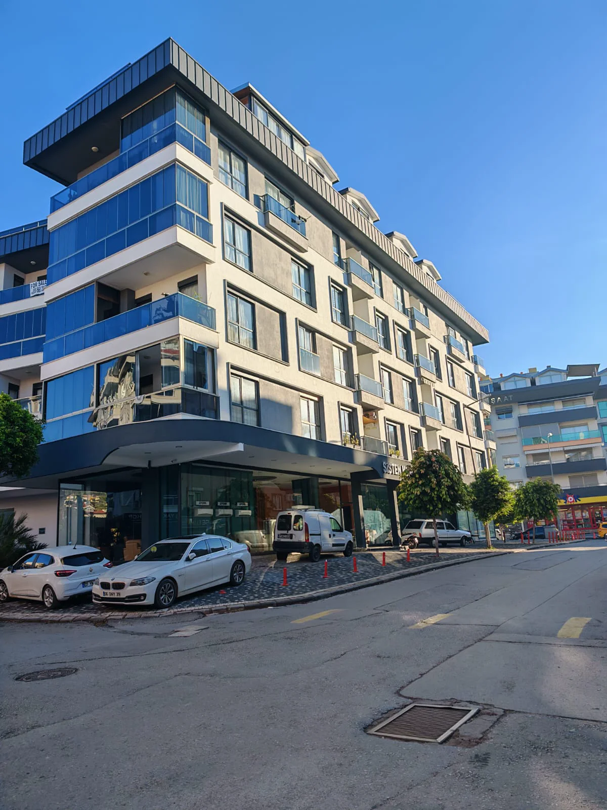 Moderne Wohnhausfassade mit Glasbalkonen. Ansicht eines modernen Wohngebäudes in städtischer Umgebung mit eleganter grau-weißer Fassade, zahlreichen Glasbalkonen und einem Eingangsbereich auf Straßenebene mit parkenden Autos.