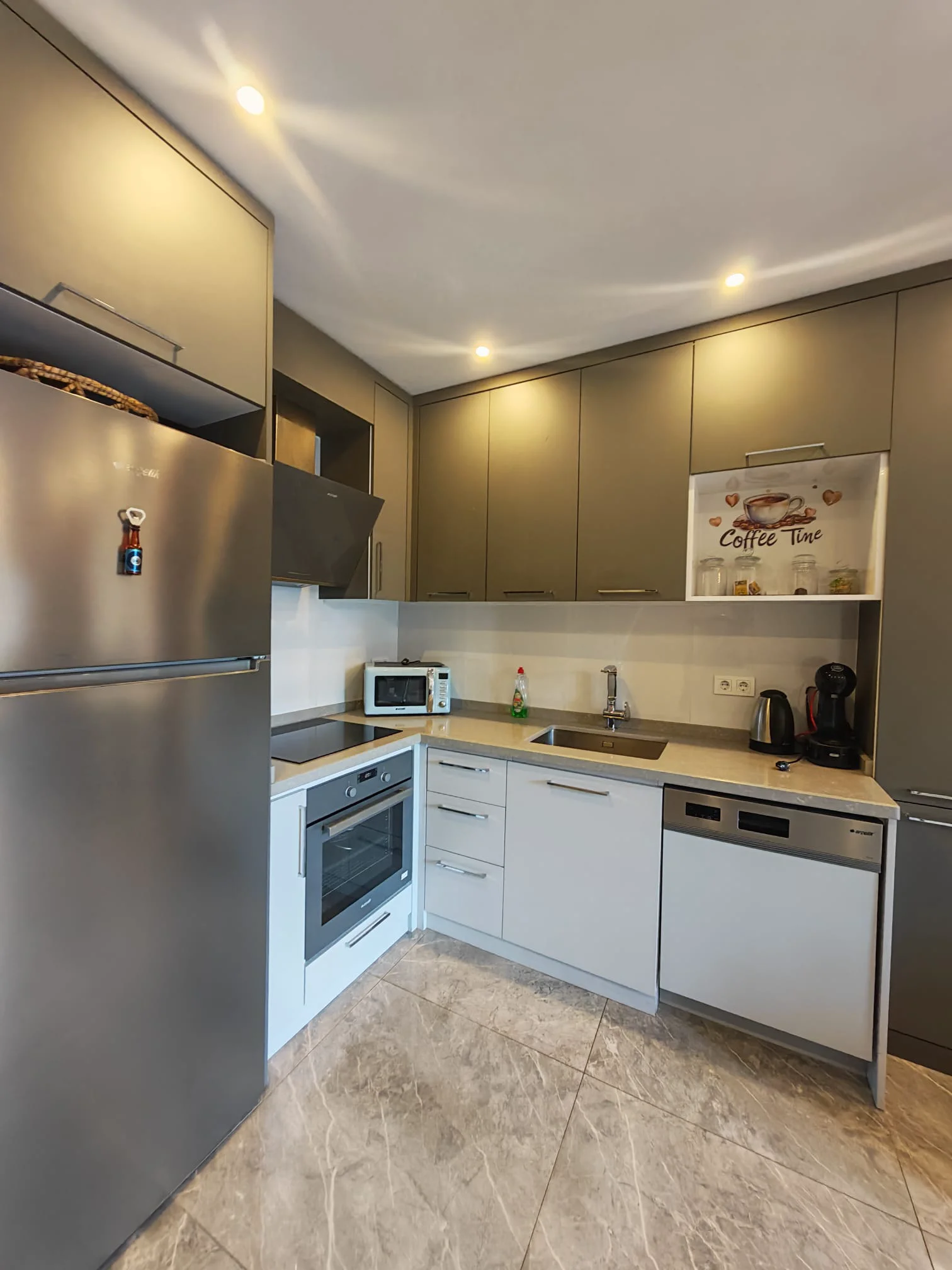 Moderne Küche mit Einbaugeräten und Fliesenboden. Eine moderne, funktionale Küche mit Edelstahlkühlschrank, Einbaubackofen und Geschirrspüler. Ausgestattet mit eleganten grauen und weißen Schränken, einer hellgrauen Arbeitsplatte und hellem Fliesenboden.
