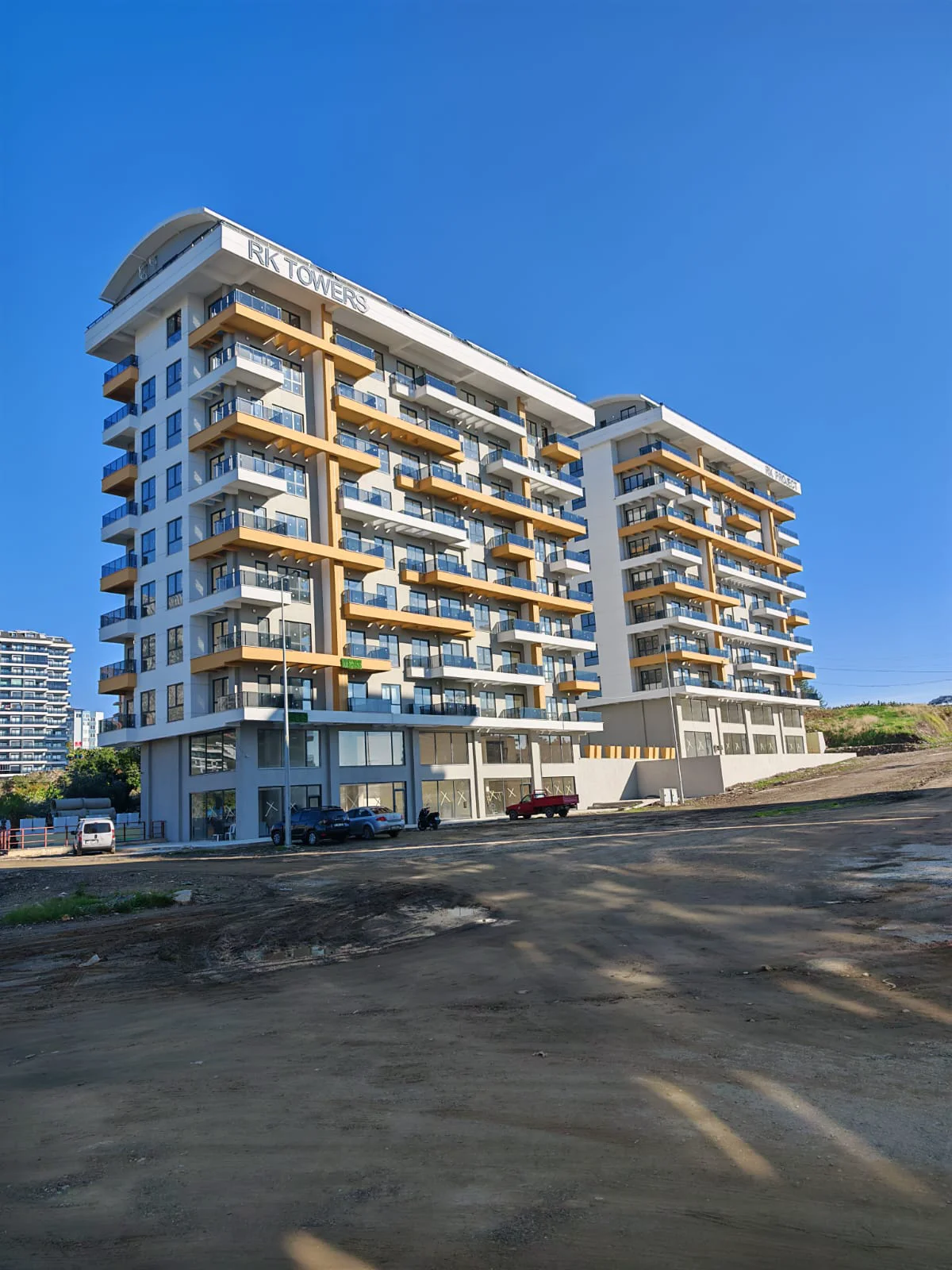 RK TOWERS Modernes Apartmentgebäude Außenansicht mit Balkonen. Außenansicht des neuen mehrstöckigen Wohngebäudes „RK TOWERS“ unter klarem blauem Himmel. Es besticht durch moderne Architektur mit zahlreichen Balkonen, Glasgeländern und Gewerbeeinheiten im Erdgeschoss.