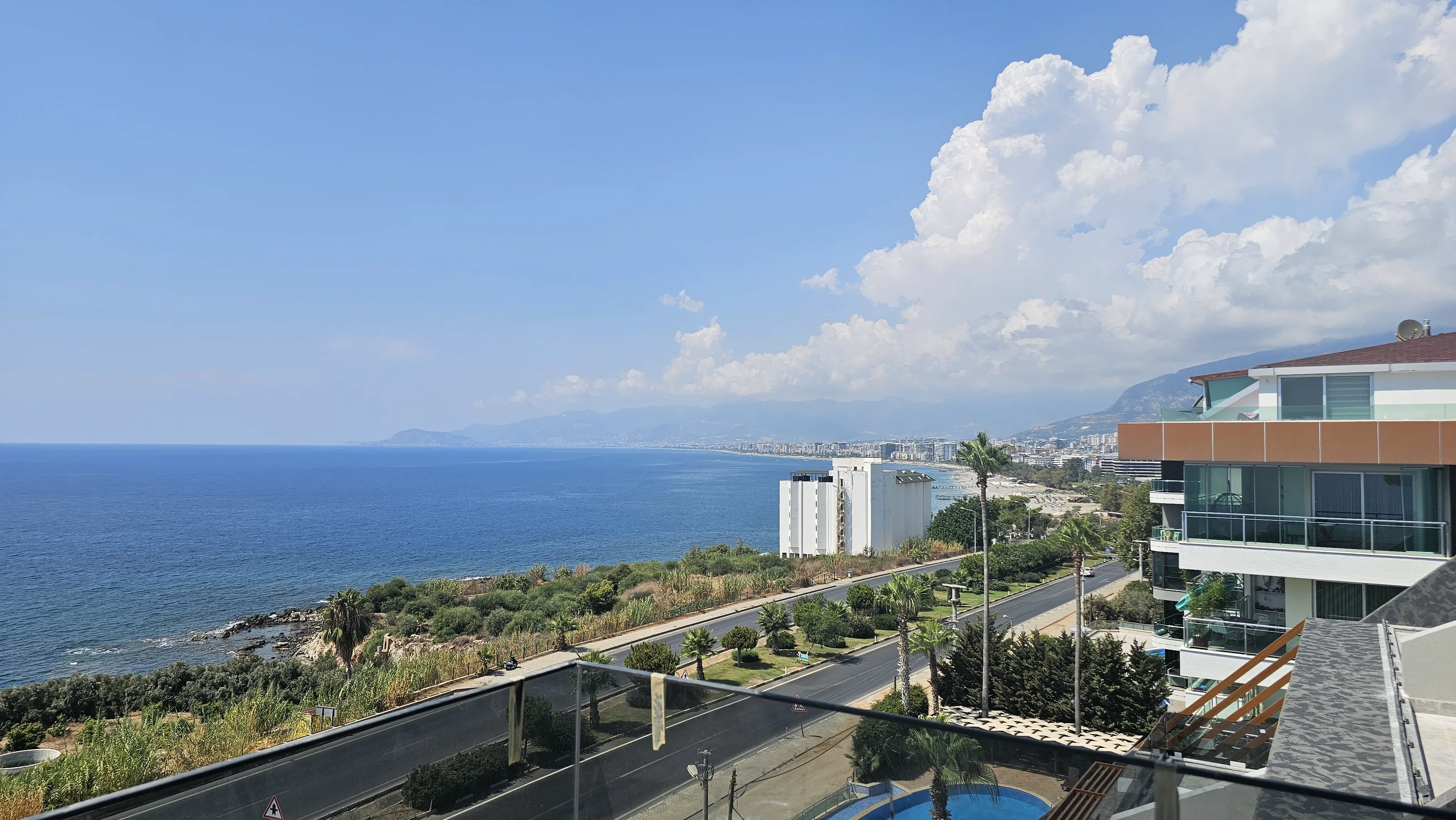 Panoramameerblick vom modernen Küstenbalkon. Ein lebhafter Ausblick im Freien von einem modernen Balkon, der das weite Mittelmeer, eine entfernte Stadt und üppiges Küstengrün unter einem klaren blauen Himmel zeigt.