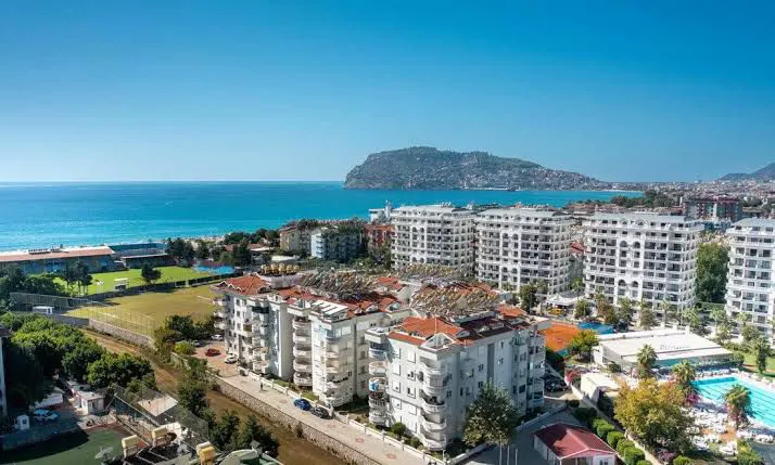 Alanya Küstenapartments mit atemberaubendem Mittelmeerblick. Luftaufnahme eines modernen Wohnkomplexes in Alanya, Türkei. Zeigt weiße Apartmentgebäude, einen Außenpool, Grünflächen und einen atemberaubenden türkisfarbenen Mittelmeerblick mit einer entfernten Insel.