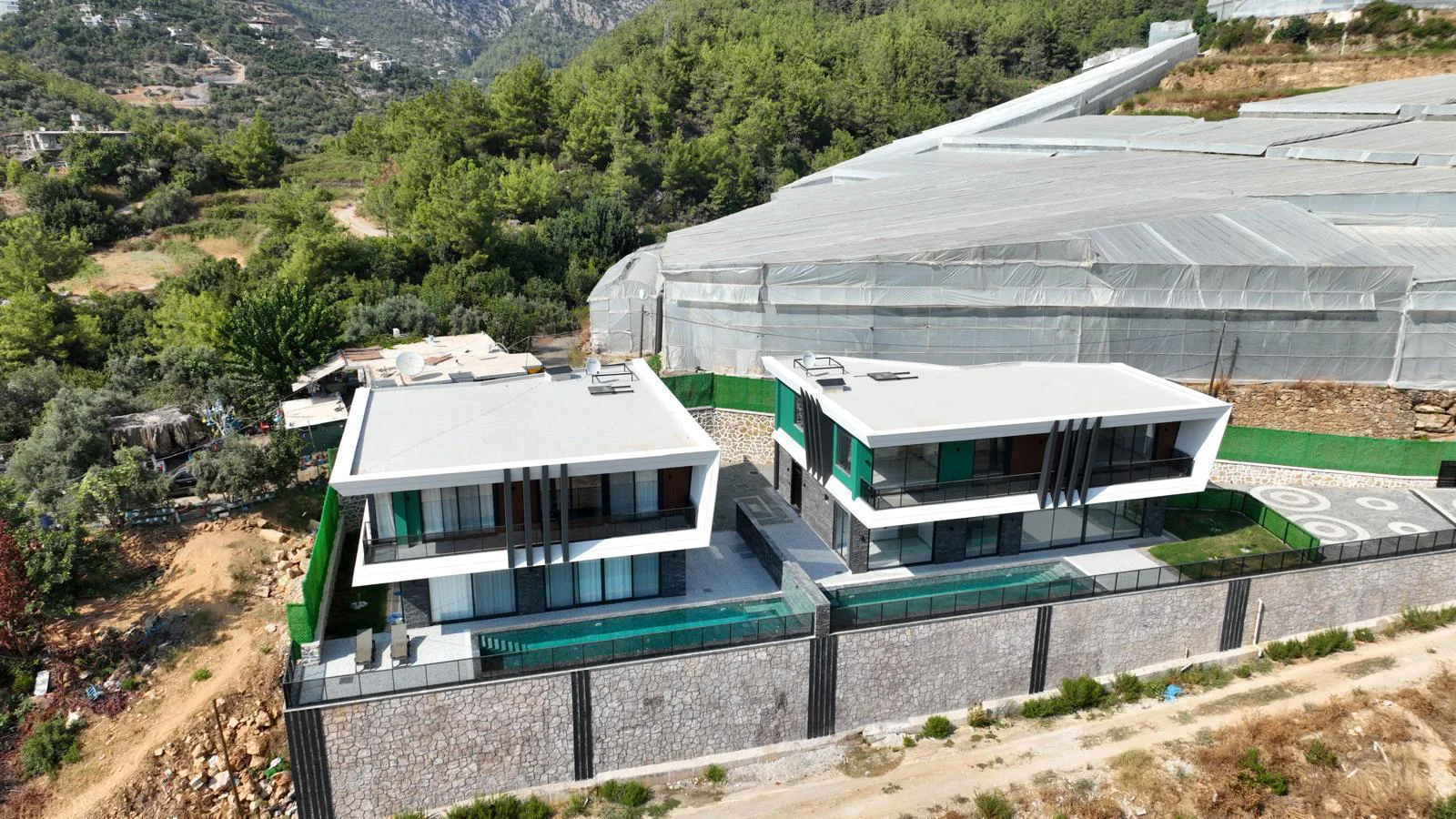Moderne Villen mit privaten Pools und Bergblick. Luftaufnahme von zwei modernen Villen mit Flachdächern und privaten Swimmingpools. Diese zeitgenössischen Häuser liegen an einem grünen Hang mit malerischem Bergblick.