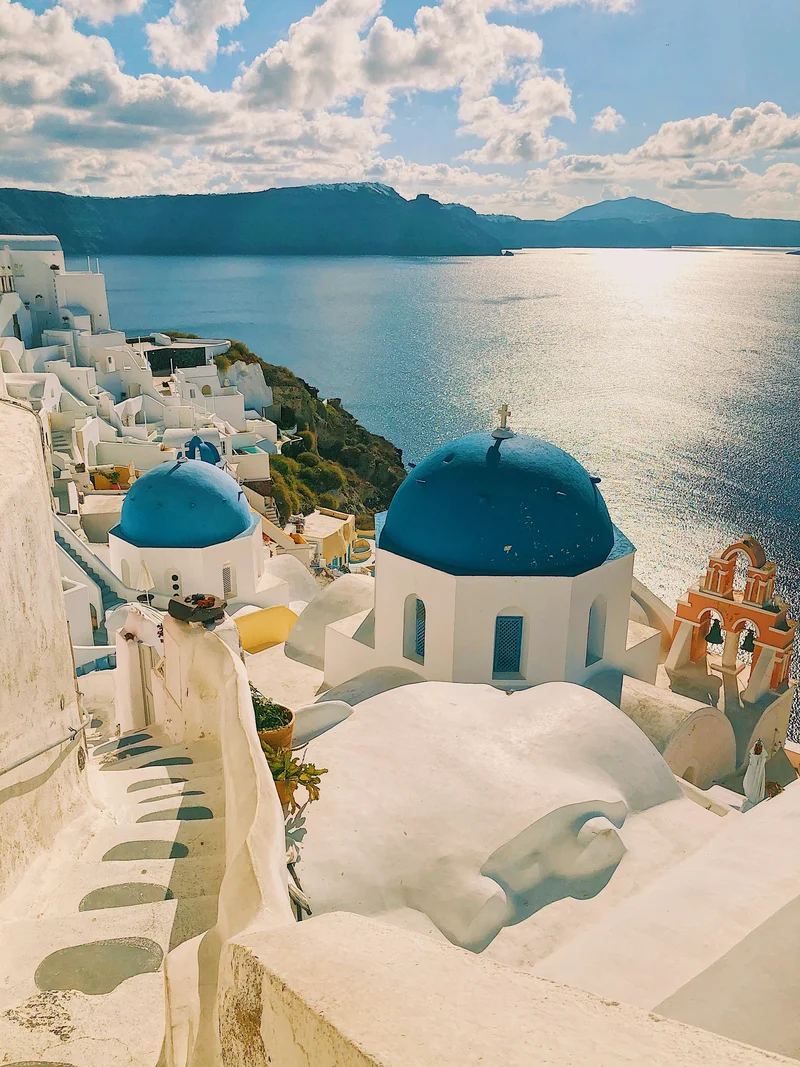 Panoramic view of a traditional white-washed coastal town with iconic blue-domed churches, overlooking the sparkling blue Aegean Sea and distant mountains under a sunny sky.