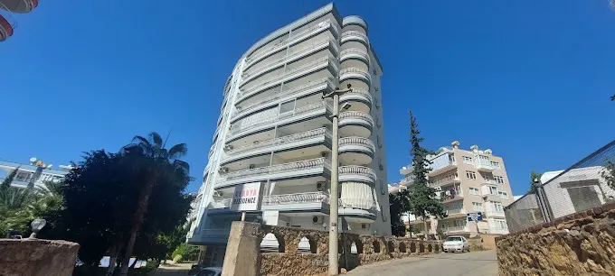 واجهة مجمع سكني حديث في ألانيا مع شرفات متعددة. منظر خارجي لمبنى سكني حديث متعدد الطوابق في ألانيا تحت سماء زرقاء صافية، يتميز بواجهة بيضاء منحنية وشرفات عديدة، يظهر من شارع محاط بأشجار النخيل.