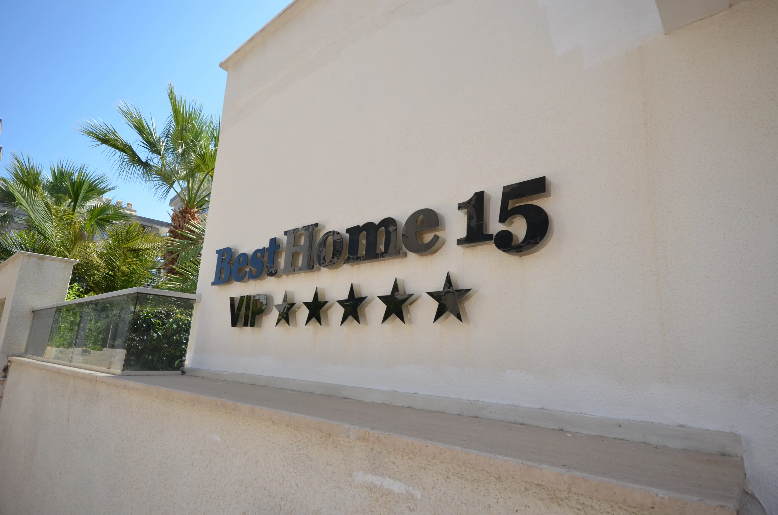 التصميم الخارجي لمبنى BestHome 15 VIP. منظر خارجي لعقار BestHome 15 VIP. يتميز بواجهة حديثة فاتحة اللون مع لافتة 'BestHome 15 VIP' البارزة ذات الخمس نجوم وأشجار نخيل مورقة تحت سماء زرقاء.